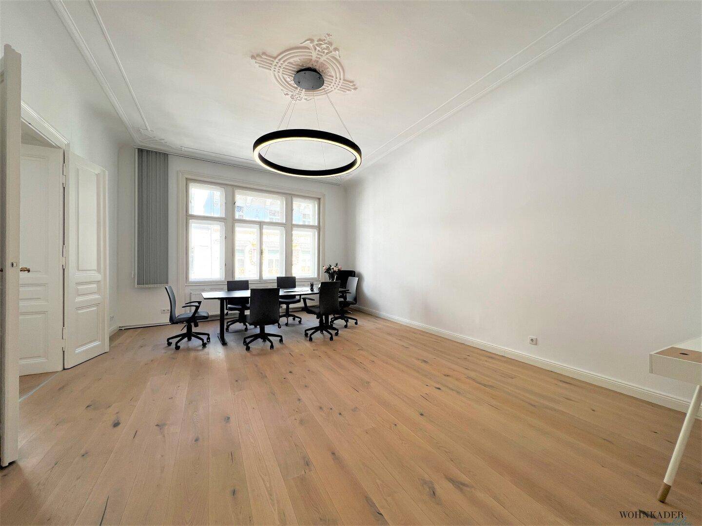 Heller Büroraum mit großem Konferenztisch, ergonomischen Stühlen und hohen Decken mit Stuckverzierungen.