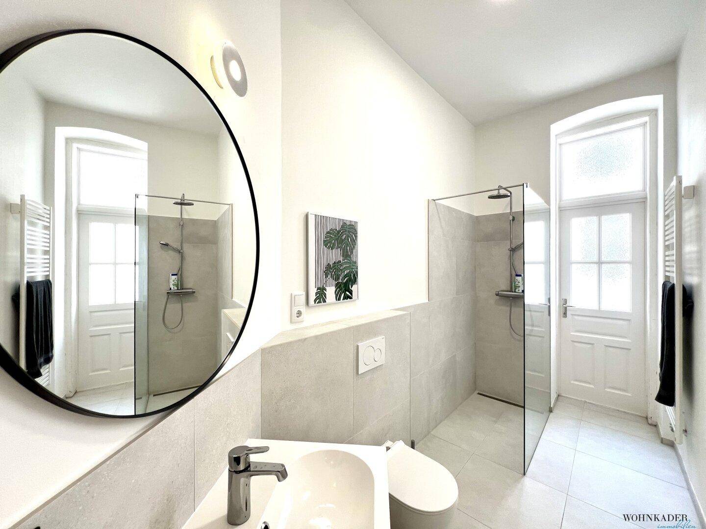 Modernes Badezimmer mit großem Rundspiegel, Waschtisch, wandhängendem WC und einer begehbaren Dusche.