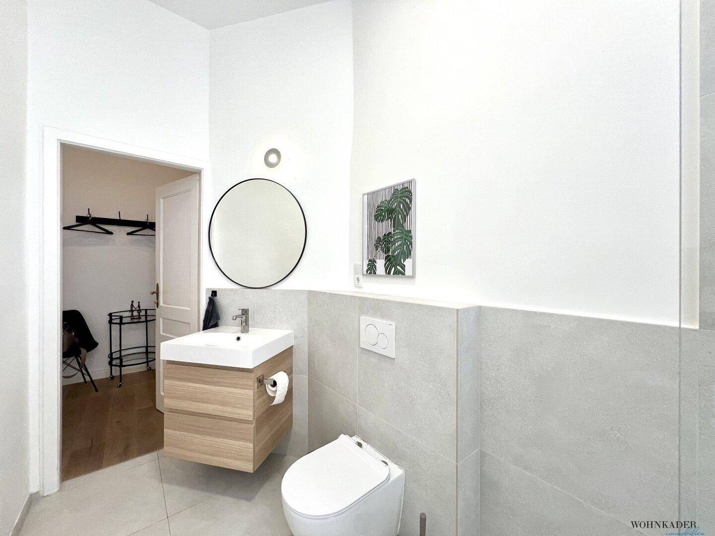 Badezimmer mit modernem Waschtisch, rundem Spiegel und wandhängendem WC, angrenzend an einen weiteren Raum.