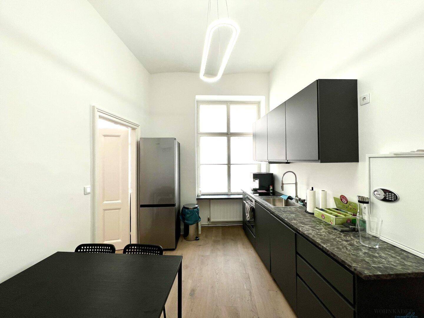 Kompakte Küche mit dunklen Unterschränken, Spüle und Kaffeemaschine, gut beleuchtet durch ein Fenster.