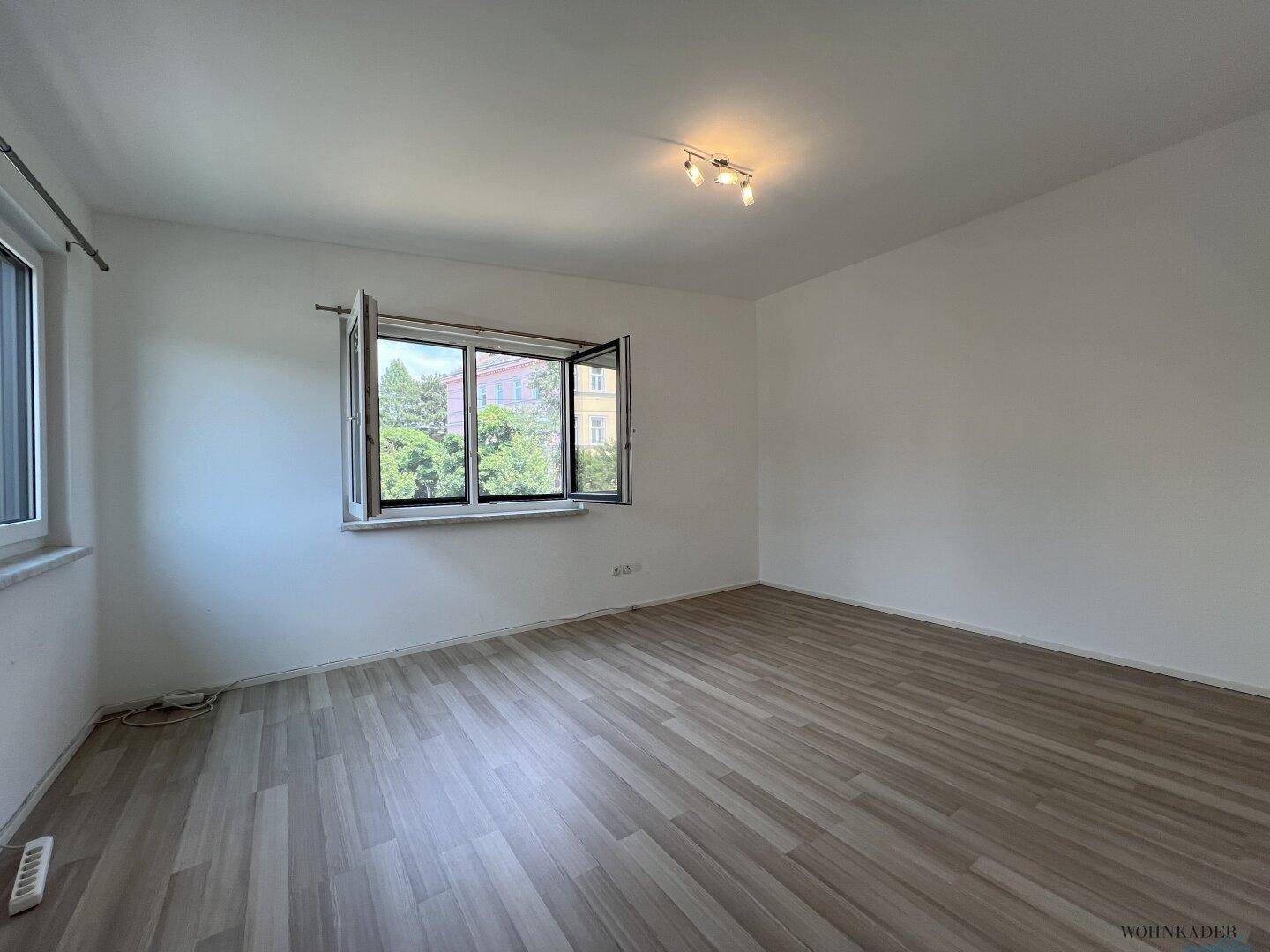 Geräumiger, heller Raum mit zwei Fenstern und hellem Laminatboden, bietet vielseitige Nutzungsmöglichkeiten.