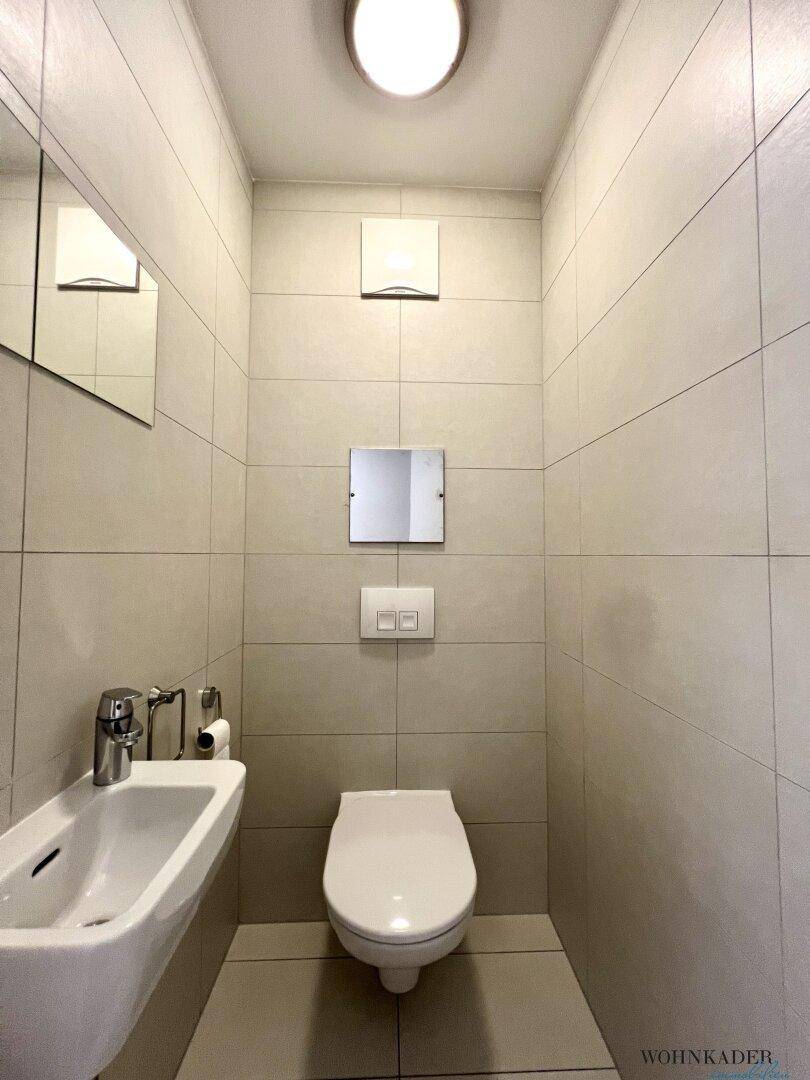 Modernes Gäste-WC mit hellen Wandfliesen, wandhängendem WC und kleinem Waschbecken.