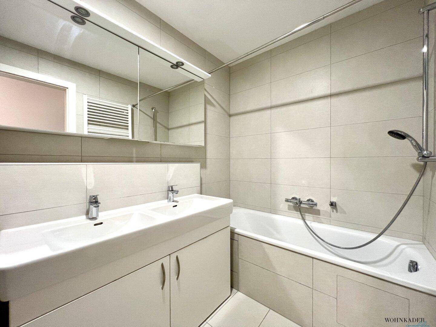 Modernes Badezimmer mit Doppelwaschbecken, Badewanne und hellen Wand- und Bodenfliesen.