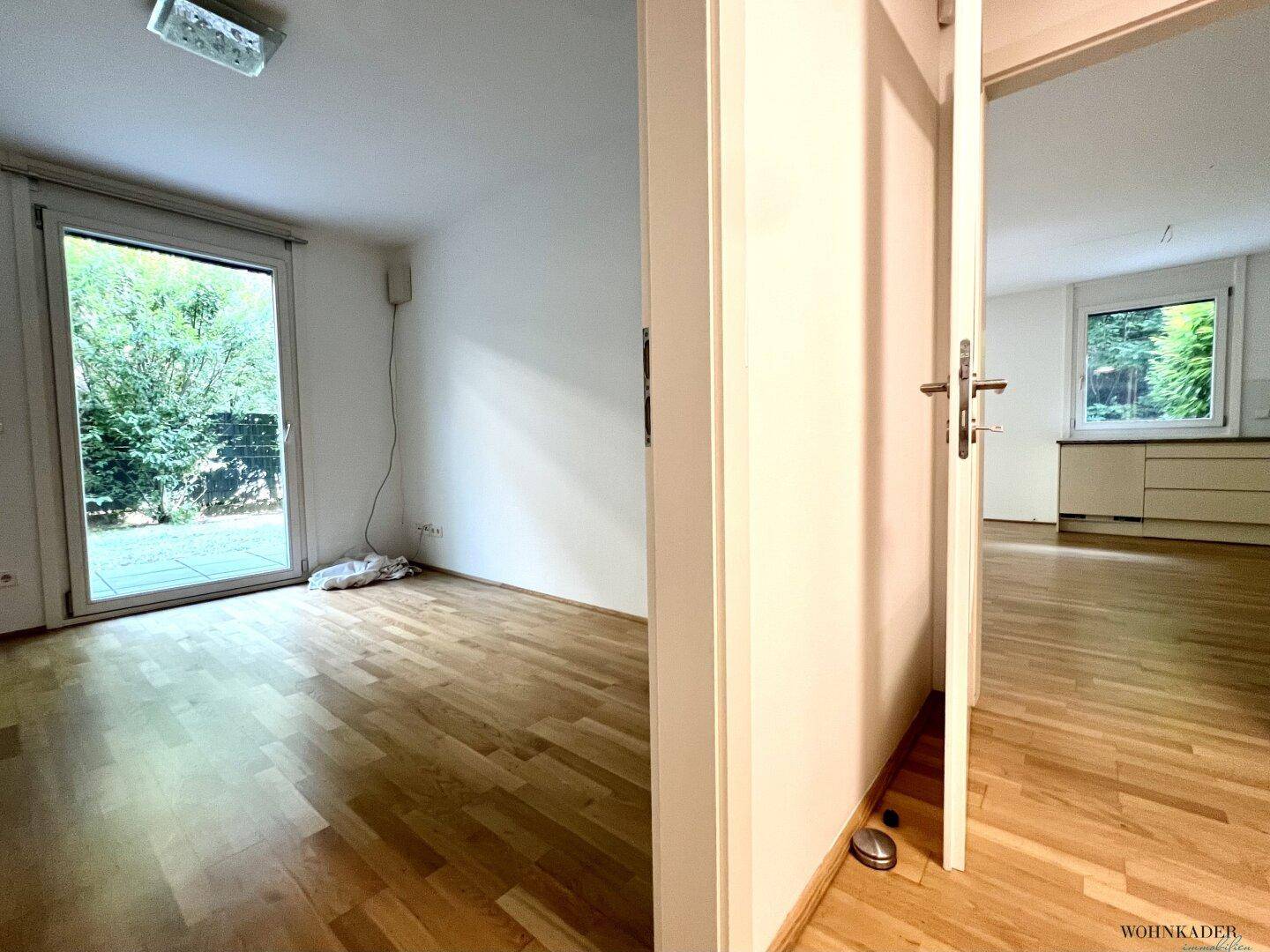 Heller Raum mit Parkettboden und Zugang zu einem weiteren Zimmer mit Blick ins Grüne.