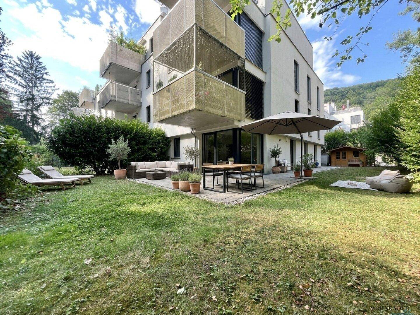 Gepflegter Garten mit Rasenfläche, möblierter Terrasse und modernem Mehrfamilienhaus im Hintergrund.