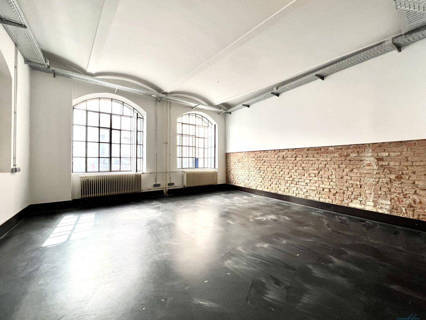 Heller, offener Raum mit hohen Decken, großen Fenstern und einer unverputzten Ziegelwand.