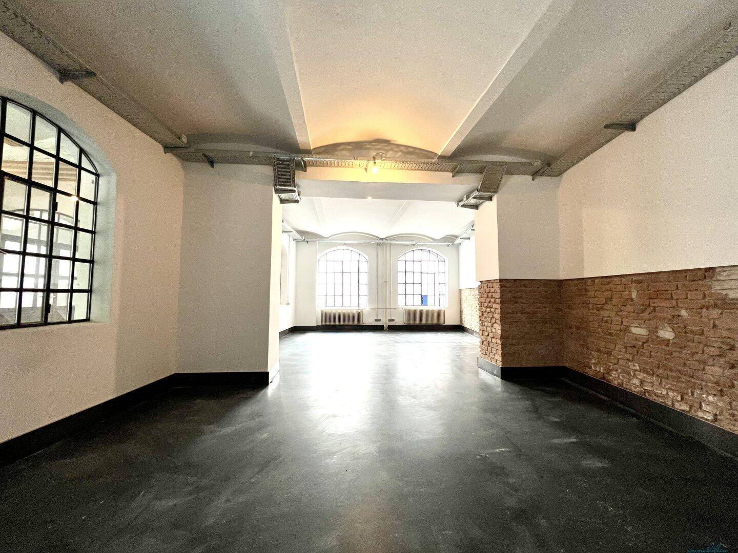 Weitläufiger, heller Raum mit hohen Decken, großen Bogenfenstern und dunklem Boden.