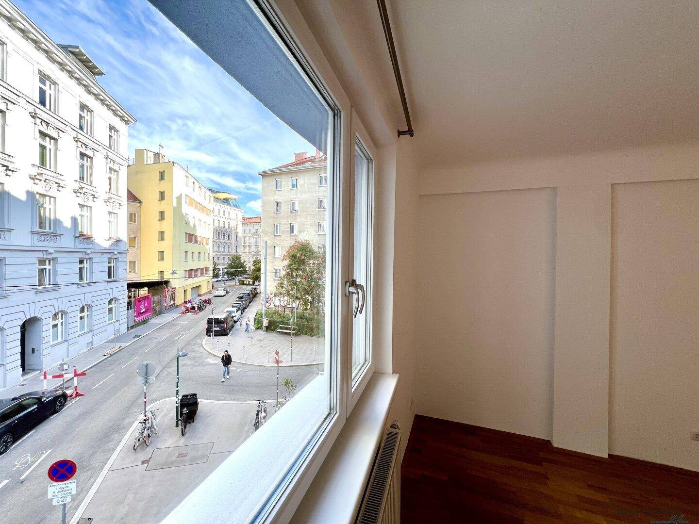 Blick aus dem Fenster auf eine belebte Stadtstraße mit historischen Gebäuden und Bäumen.