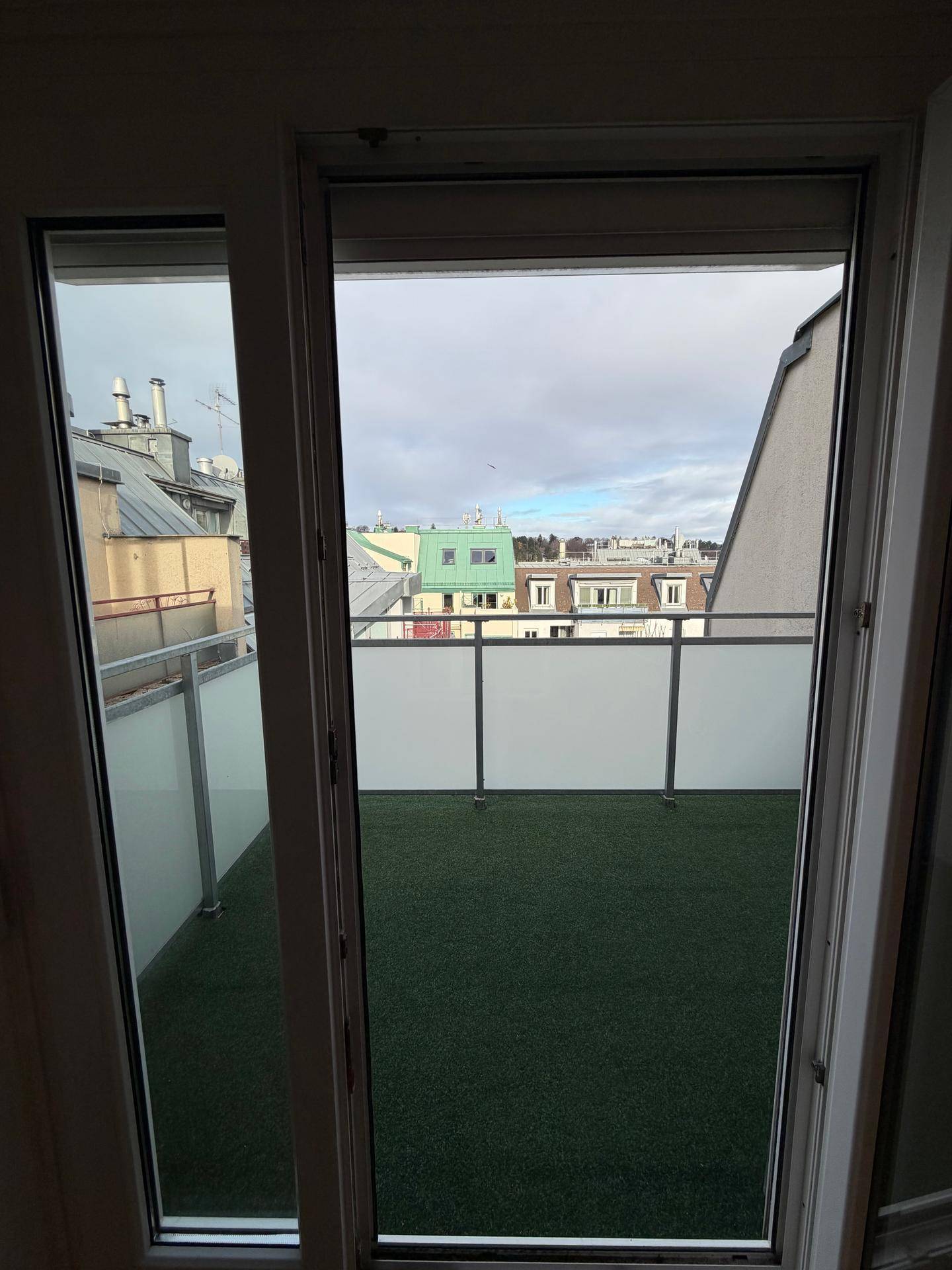 Blick vom Wohnzimmer durch die Balkontür auf den Balkon mit Kunstrasen und die Stadtkulisse.
