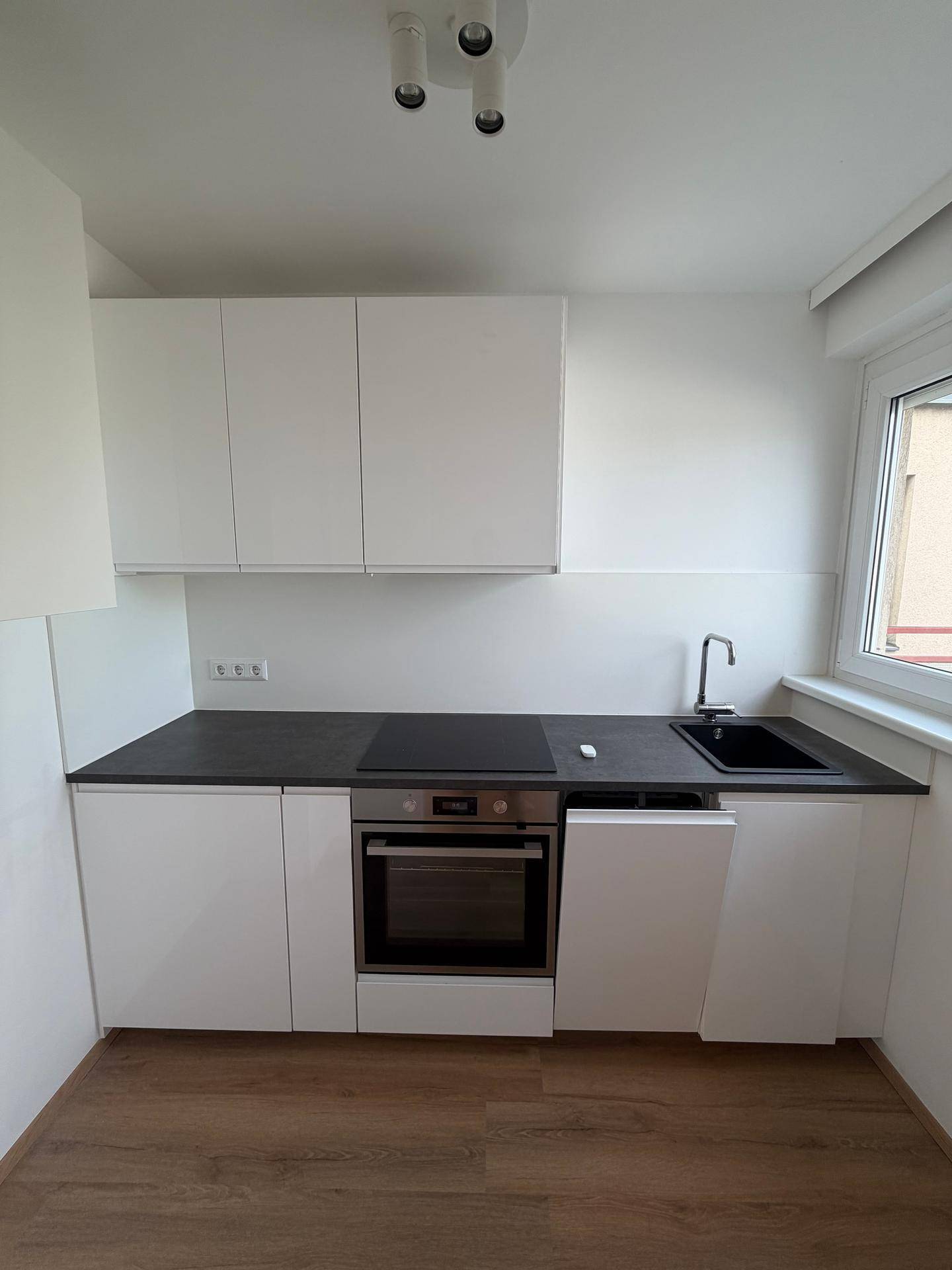 Moderne Küchenzeile mit weißen Schränken, dunkler Arbeitsplatte und integrierten Geräten am Fenster.
