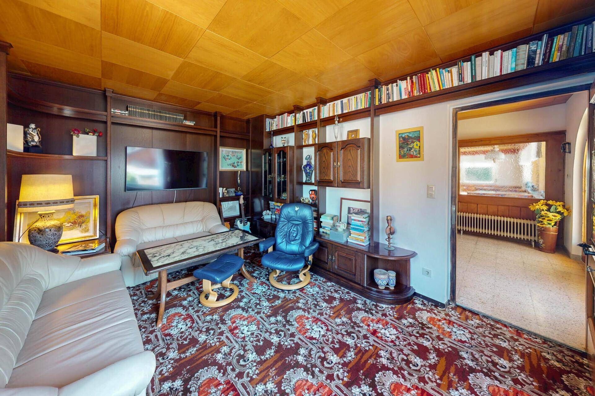 Gemütliches Wohnzimmer mit dunklen Holzmöbeln, Teppichboden und Bücherregalen.