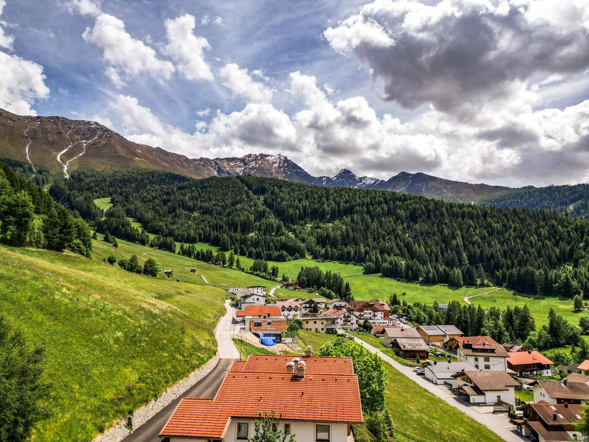 Panoramablick auf ein malerisches Dorf in einem Tal, umgeben von Wäldern und hohen Bergen.