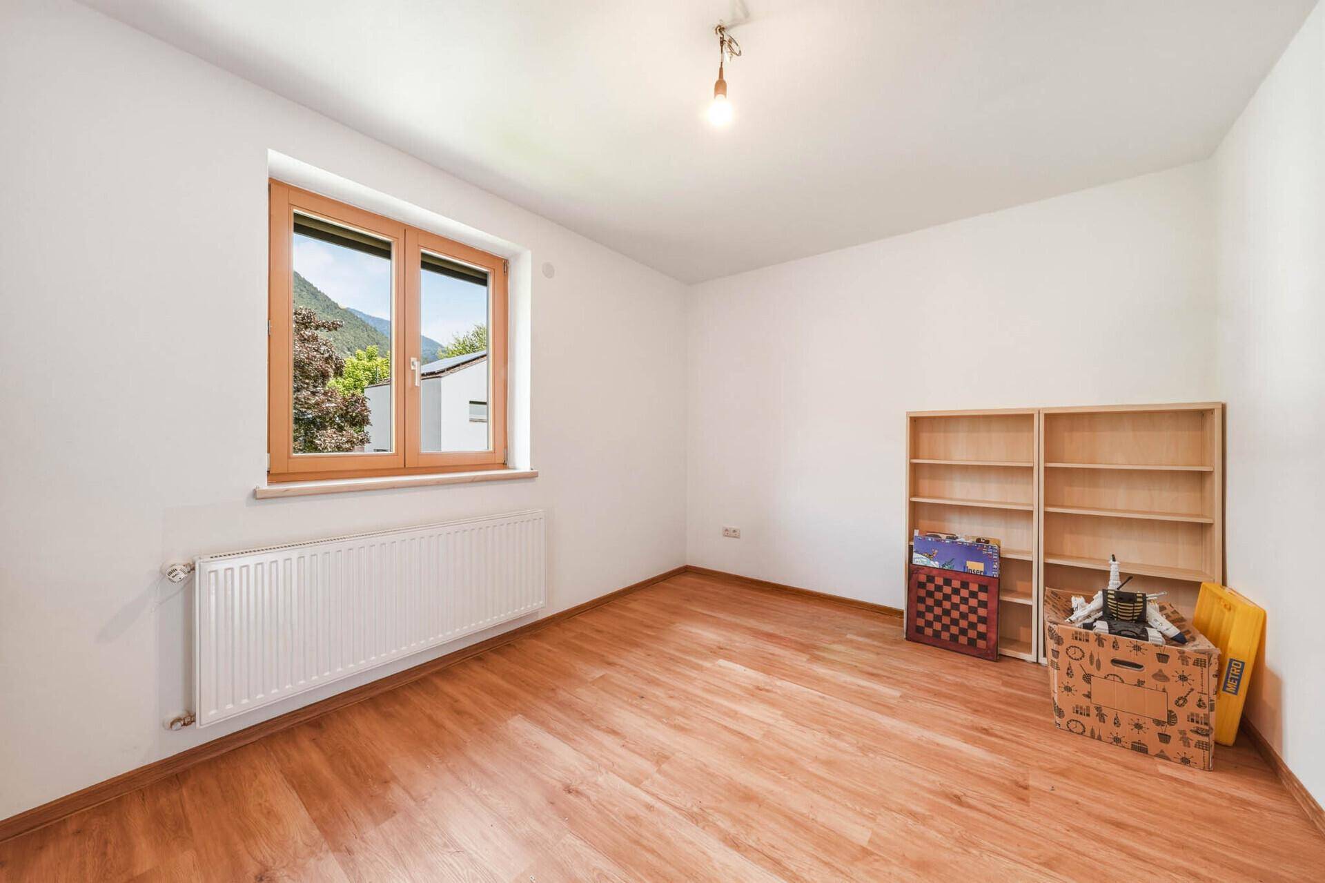 Leeres Zimmer mit Laminatboden, Heizkörper und einem Fenster mit Blick ins Grüne.