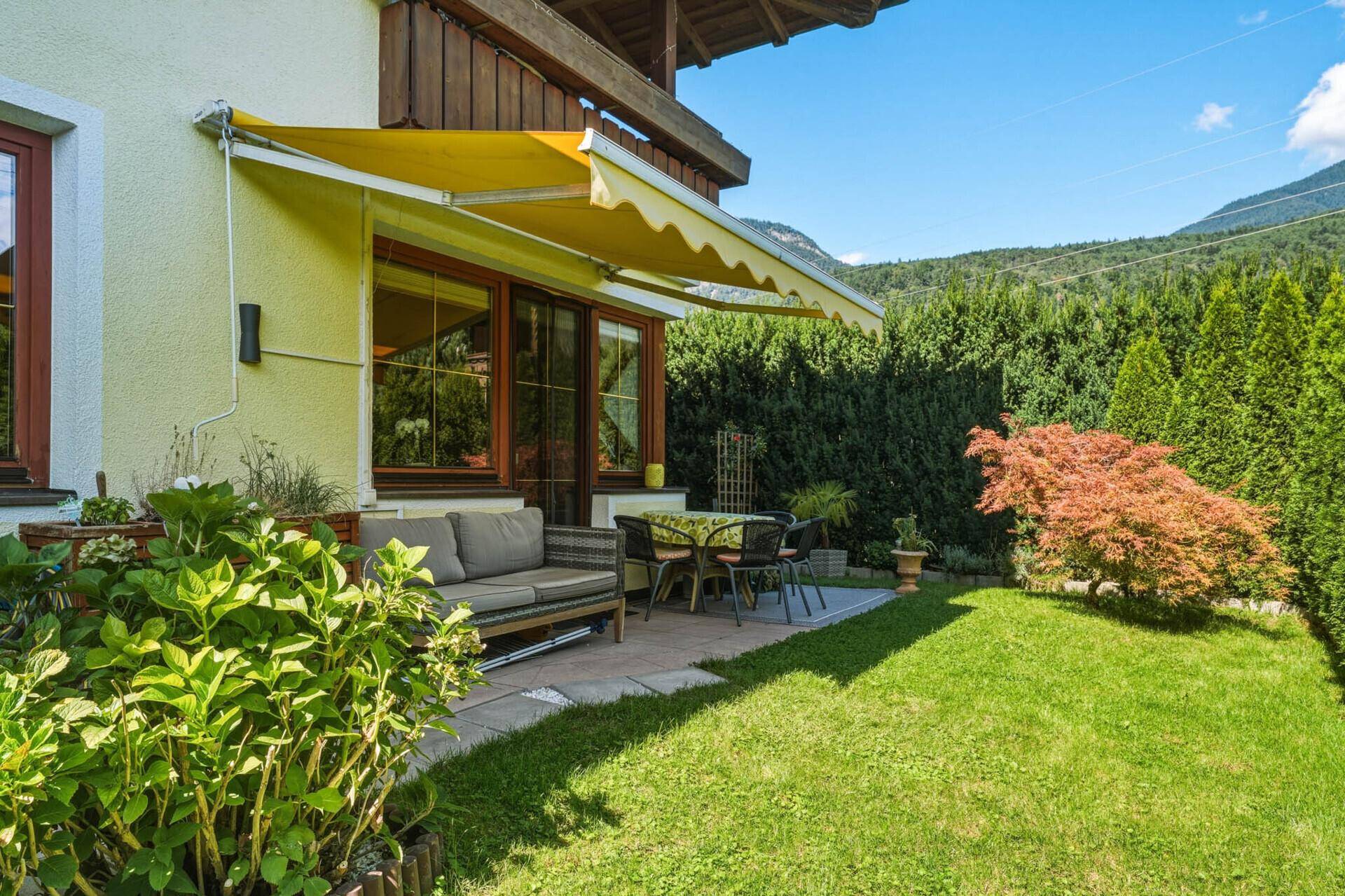Idyllische Terrasse mit Gartenmöbeln und Rasenfläche, umgeben von Hecken und Bergblick.