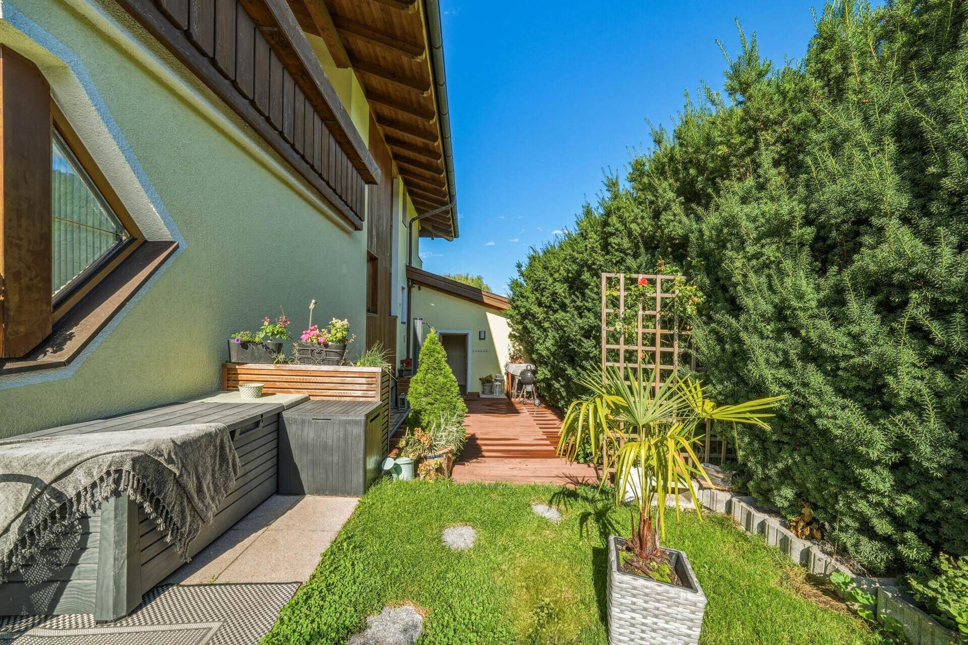 Hinterer Gartenbereich mit Holzdeckweg, hohen Hecken und dekorativen Pflanzen.