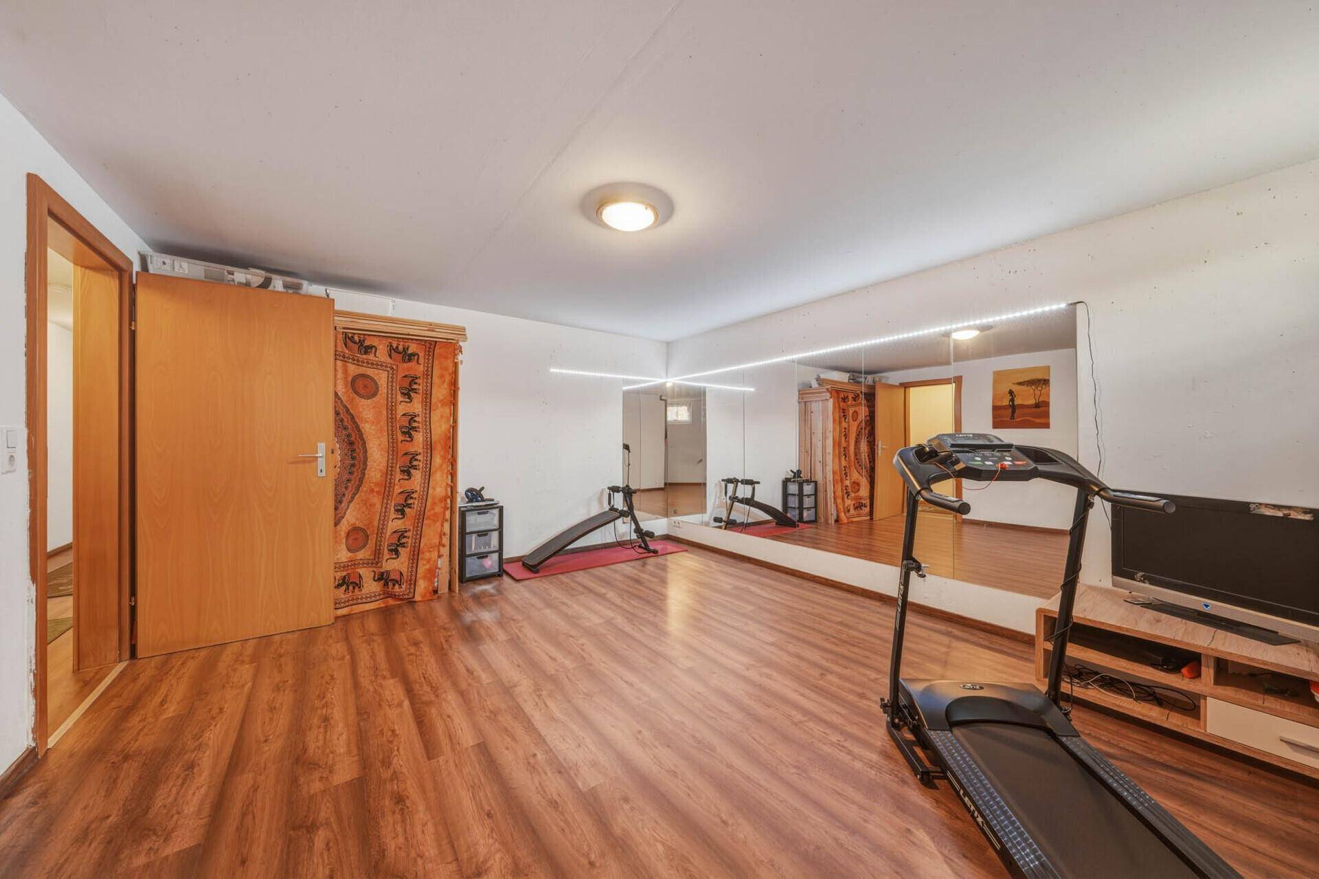 Fitnessraum im Untergeschoss mit Holzboden, großer Spiegelwand und Zugang zu anderen Räumen.