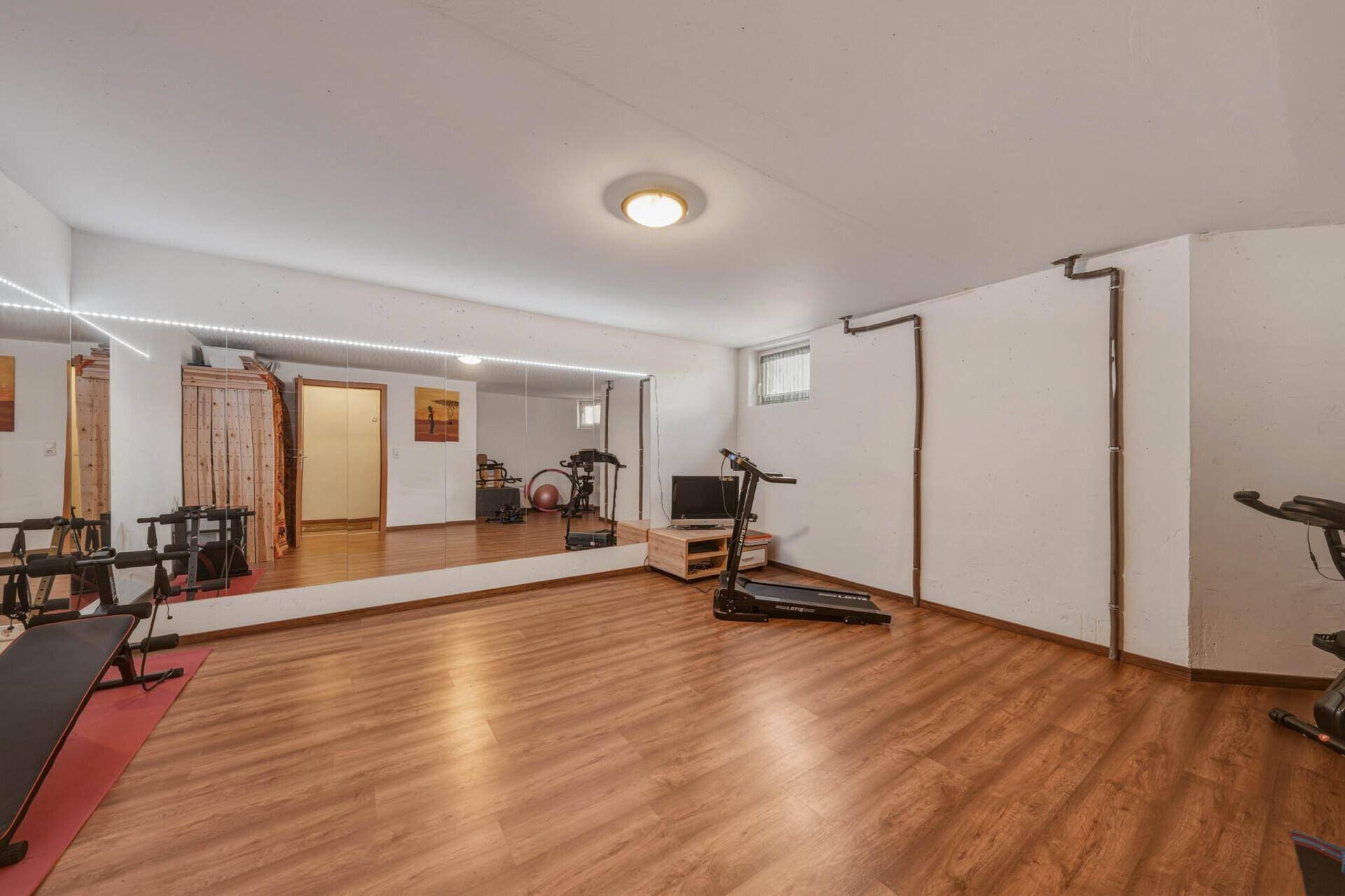 Geräumiger Fitnessraum im Untergeschoss mit Holzboden, Spiegelwand und verschiedenen Trainingsgeräten.