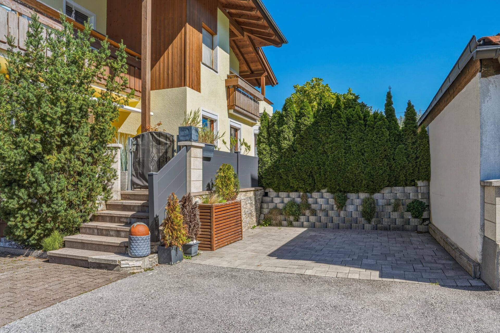 Seitenansicht des Hauses mit gepflastertem Parkplatz und einer begrünten Stützmauer.