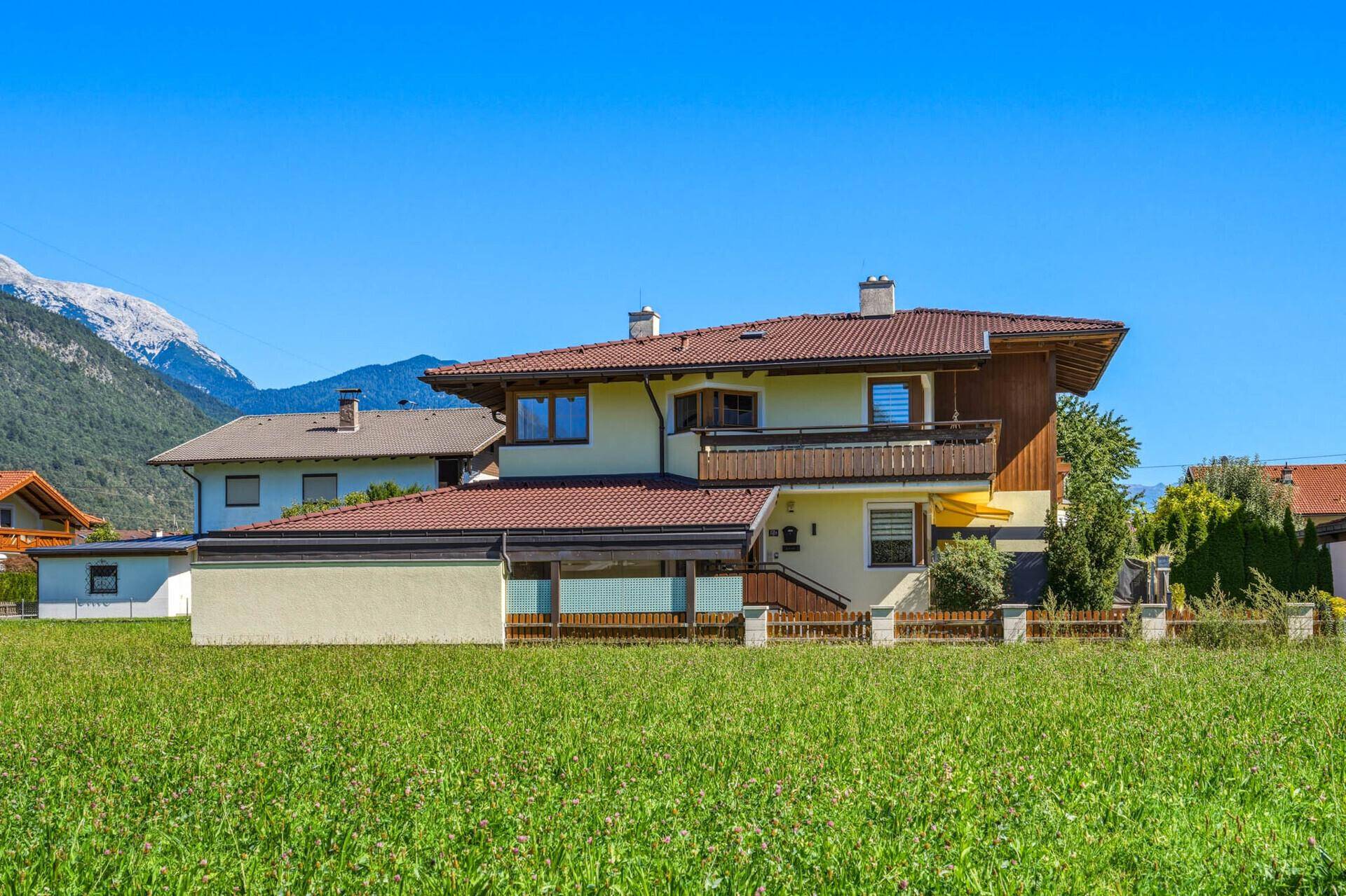Hausansicht von der Seite mit rotem Ziegeldach, umgeben von einer weiten grünen Wiese und Bergen.