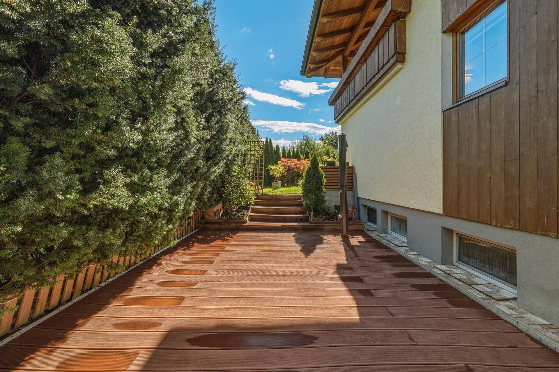 Langer Holzdeckweg entlang der Hausseite, umgeben von hohen, grünen Hecken.