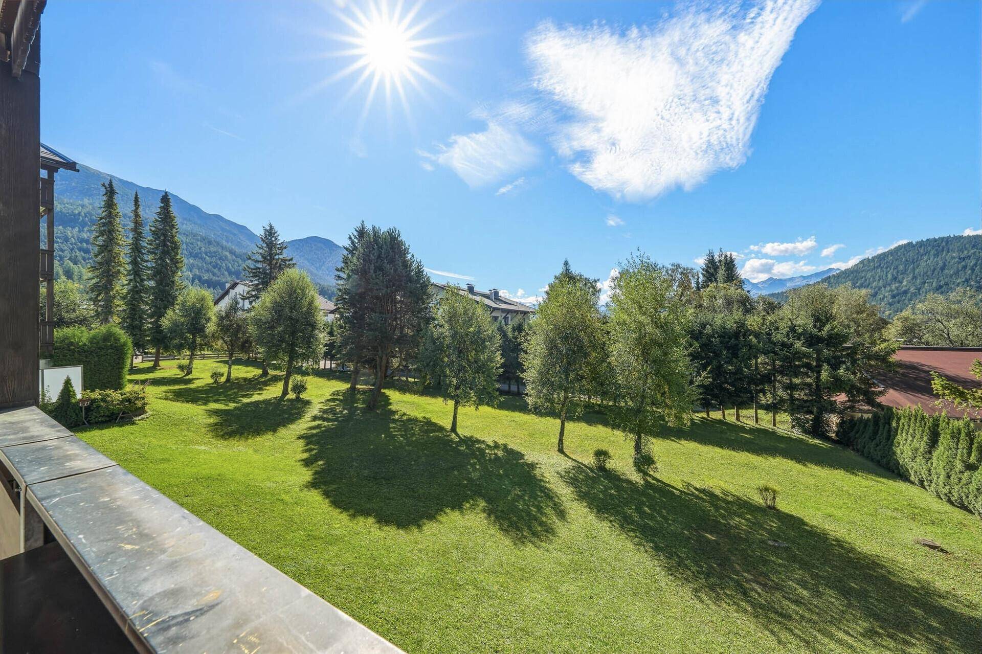Sonniger Balkon mit weitem Blick auf eine grüne Wiese und die umliegende Berglandschaft.