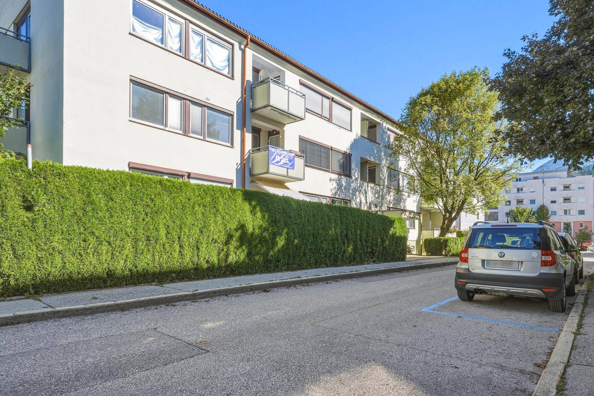Ansicht eines Wohngebäudes von der Straße mit grüner Hecke und parkenden Autos.