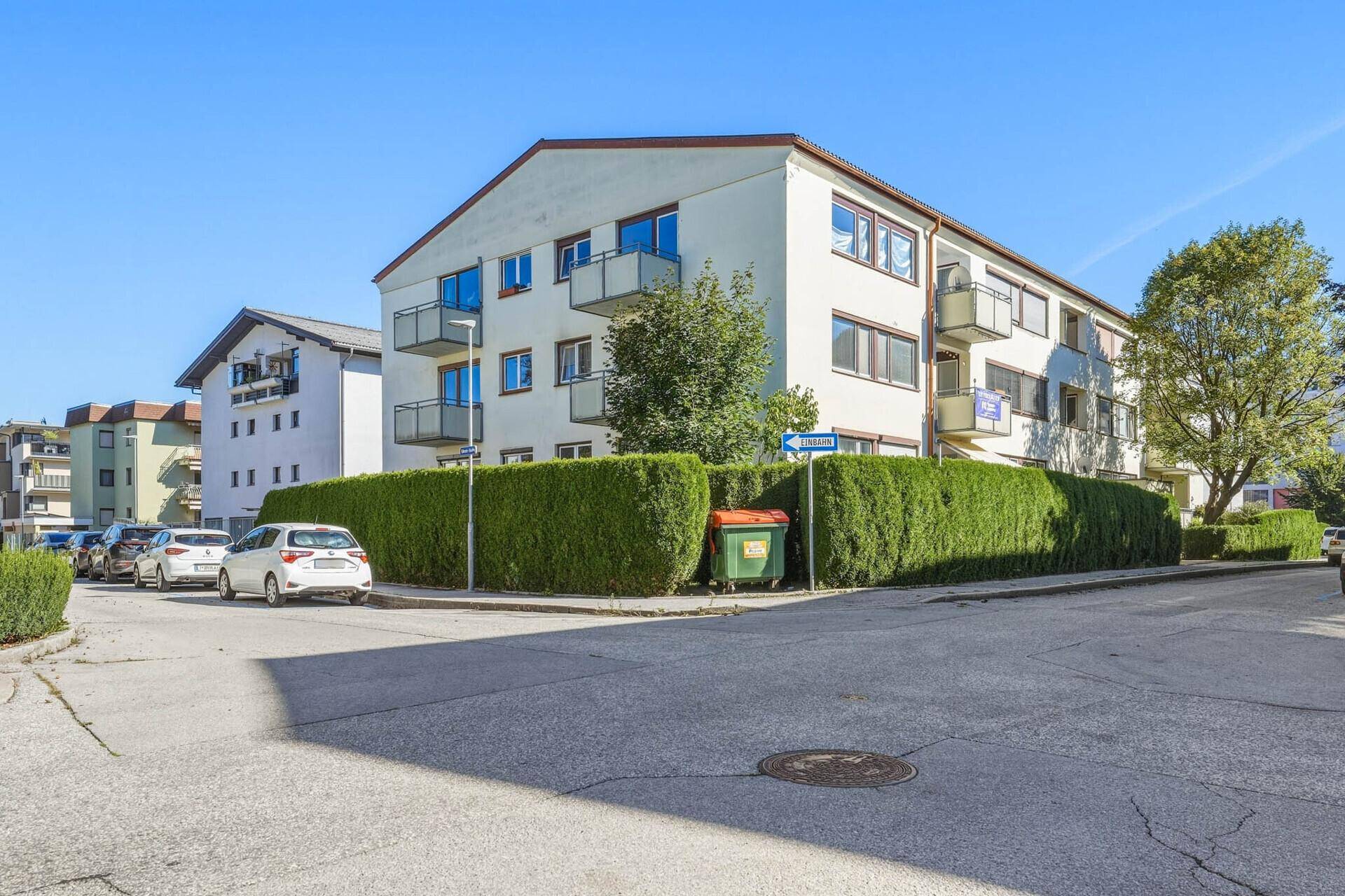 Eckansicht des Wohngebäudes von der Straße mit Hecken und Mülltonnen.