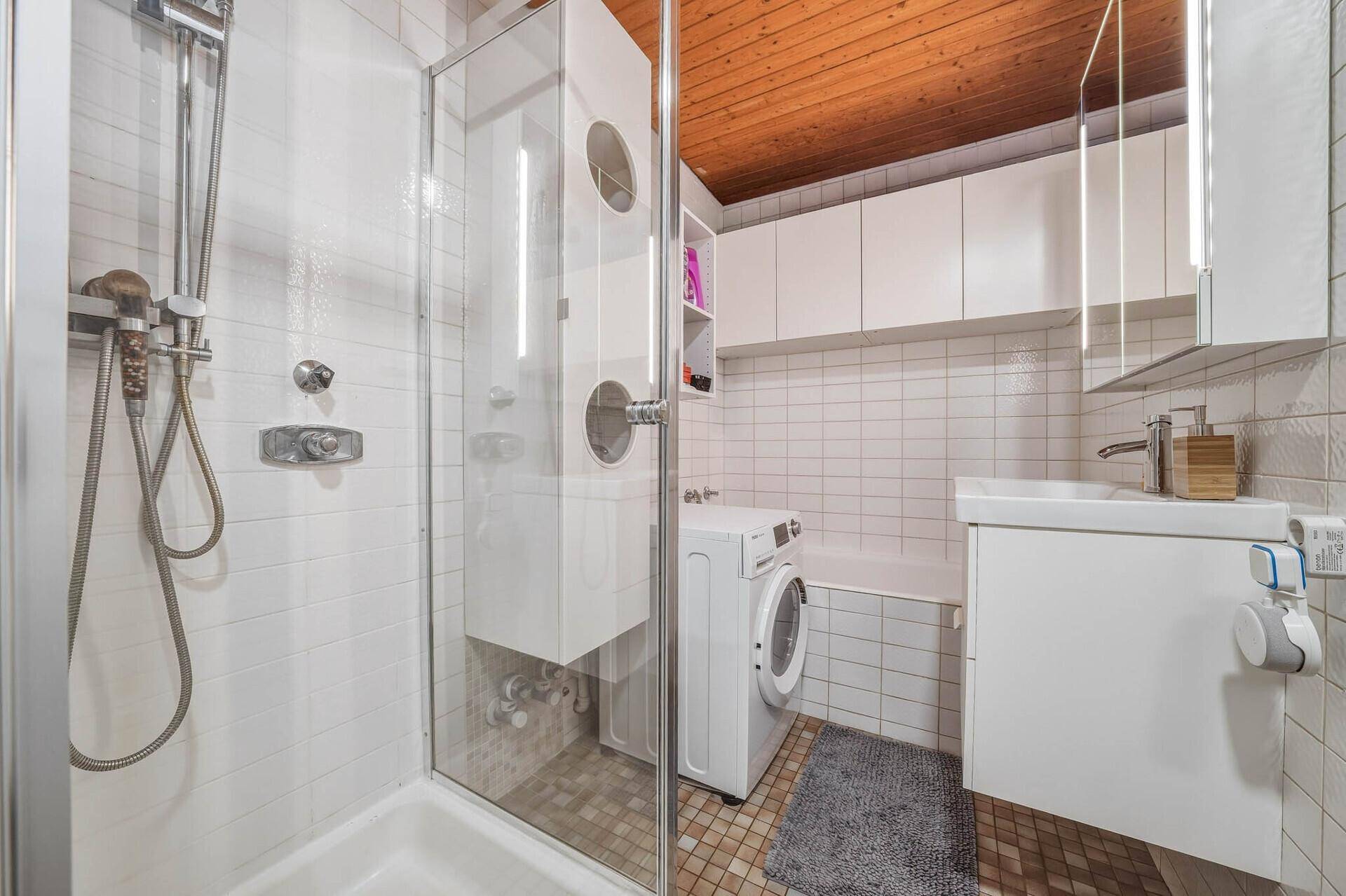 Modernes Badezimmer mit Duschkabine, Waschbecken und Waschmaschine unter Holzdecke.