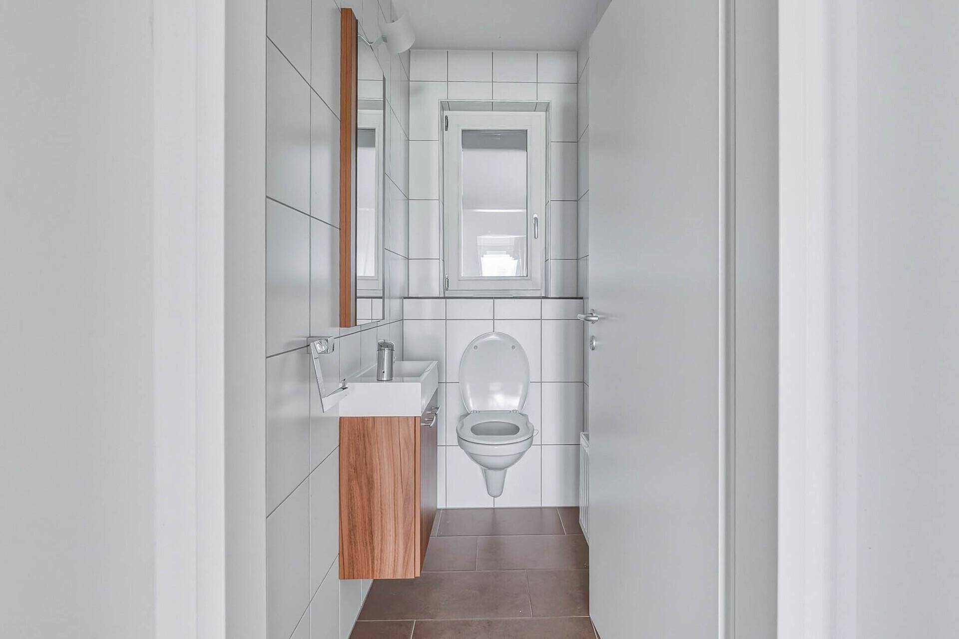 Modernes Gäste-WC mit Wand-WC, kleinem Waschbecken und Fenster, hell gefliest.