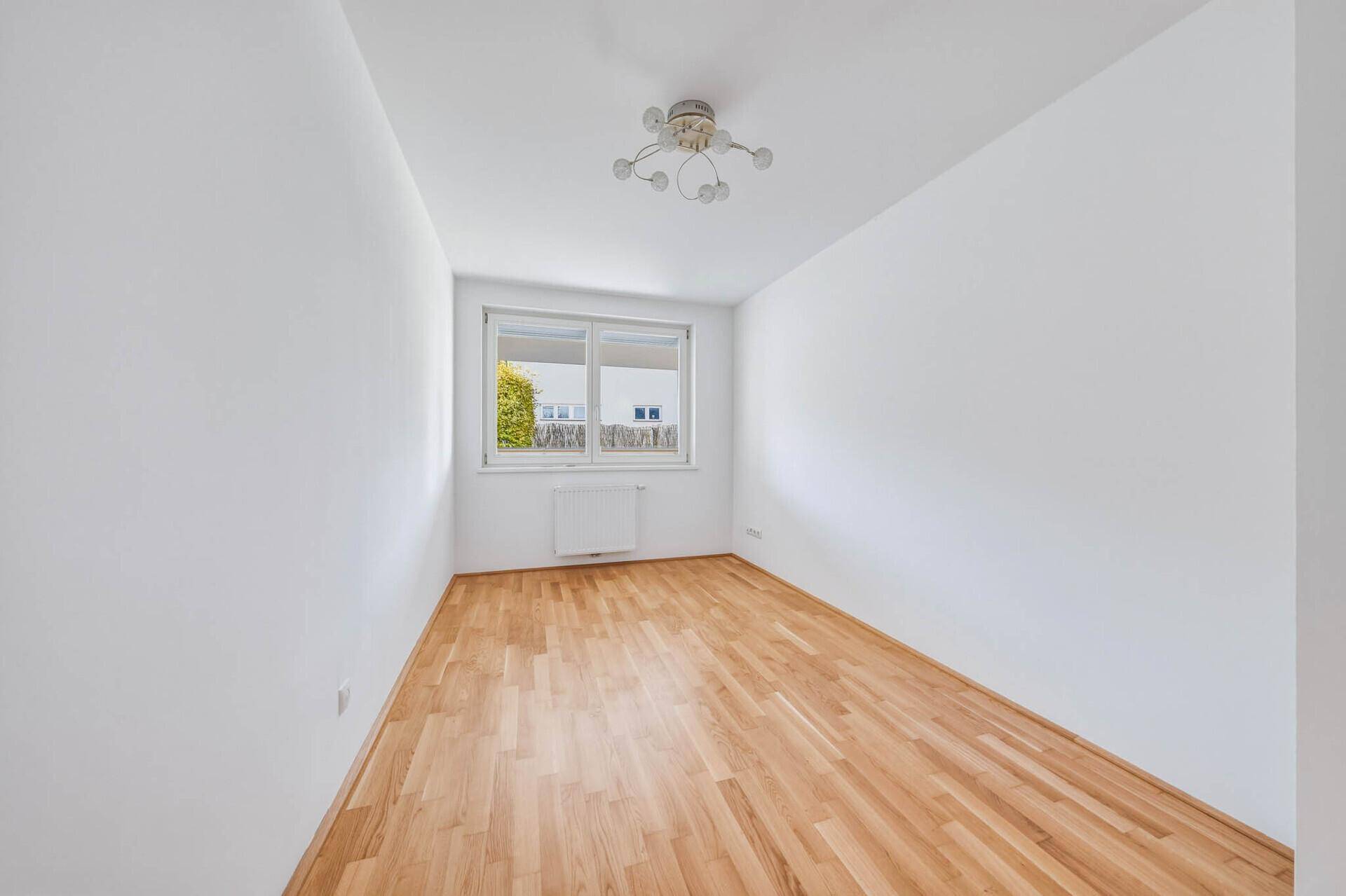 Leeres Schlafzimmer mit Parkettboden, weiß gestrichenen Wänden und einem Fenster.