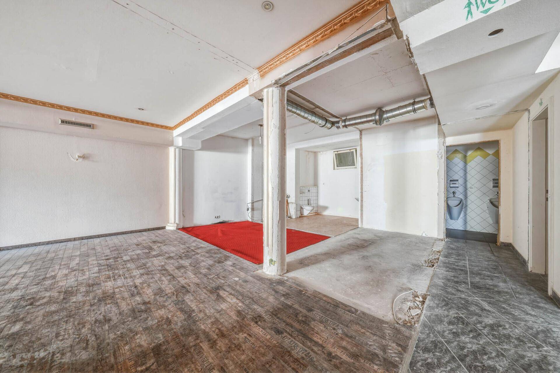 Großer Gewerberaum mit unterschiedlichen Bodenbelägen, rotem Teppich und Zugang zu Toiletten.