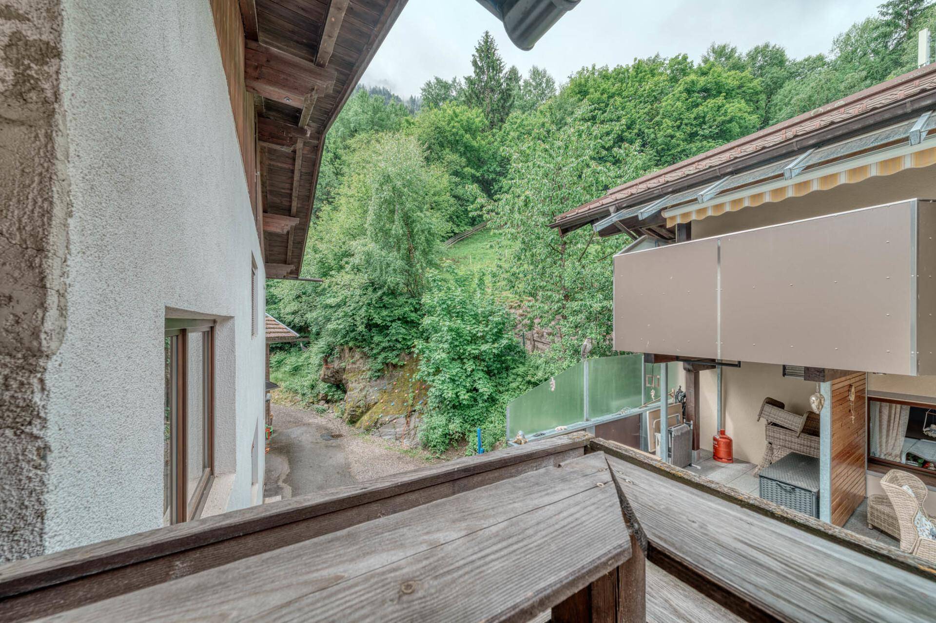 Blick von einem Balkon auf grüne Hügel, Bäume und benachbarte Gebäude in ländlicher Umgebung.