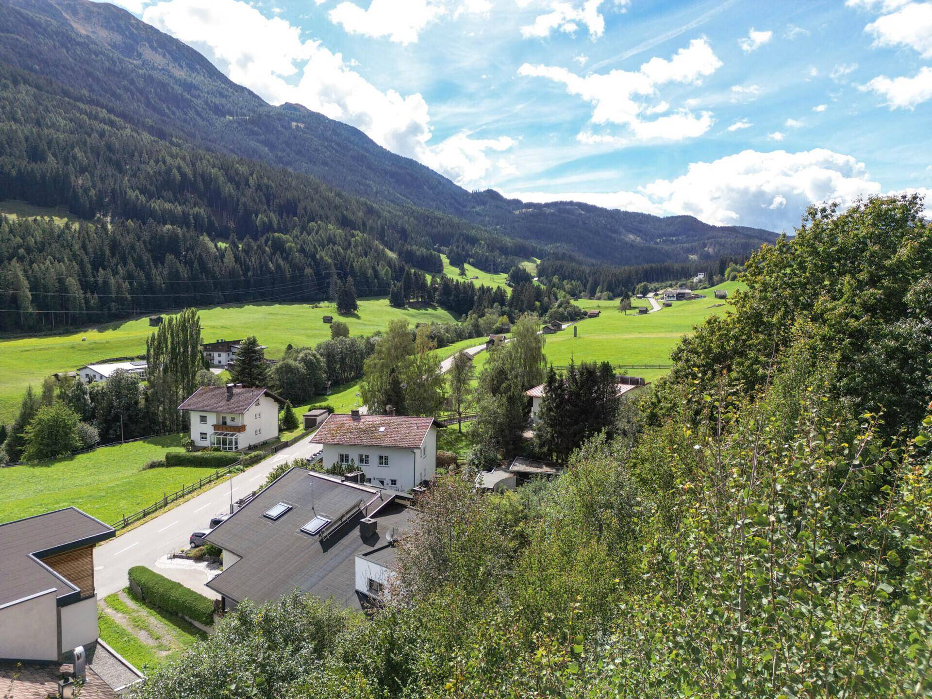 Blick über eine Ansammlung von Häusern in einem grünen Tal, umgeben von bewaldeten Bergen.