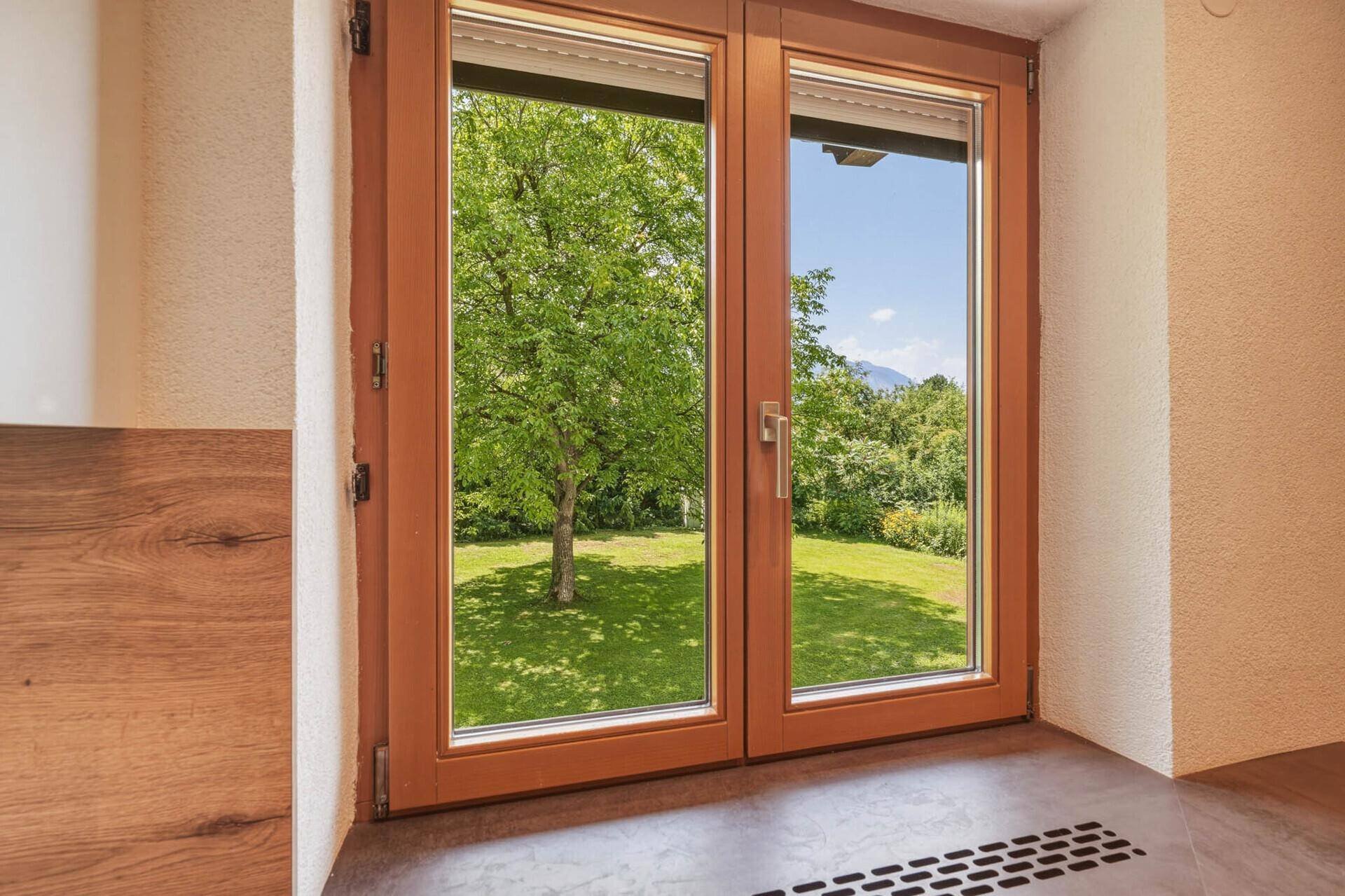 Fenster mit Blick auf den grünen Garten und die umliegende Natur an einem sonnigen Tag.
