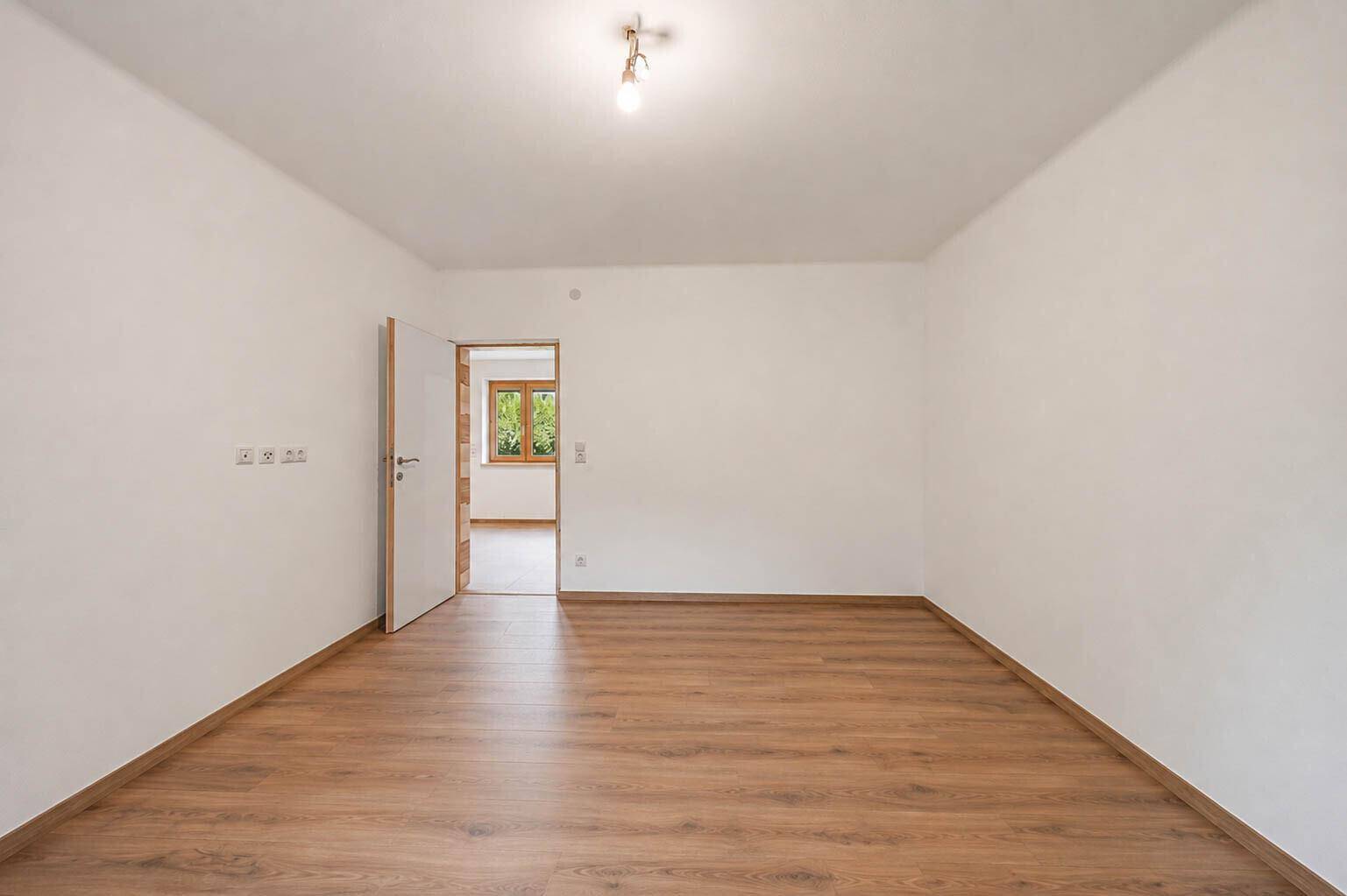 Leeres Wohnzimmer mit hellem Laminatboden, weißen Wänden und einem Fenster mit Blick ins Grüne.