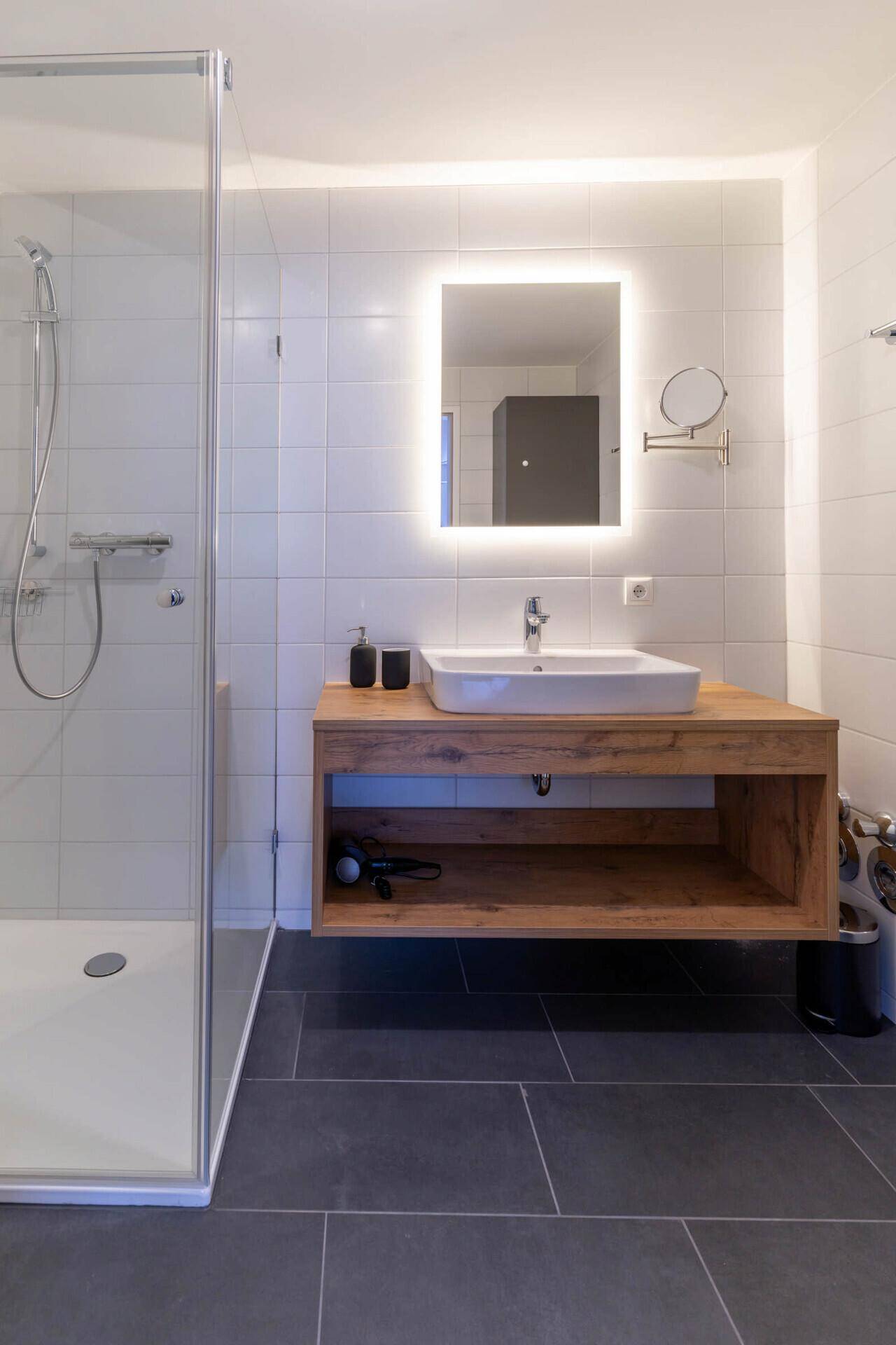 Modernes Badezimmer mit begehbarer Dusche, Holzwaschtisch und beleuchtetem Spiegel.