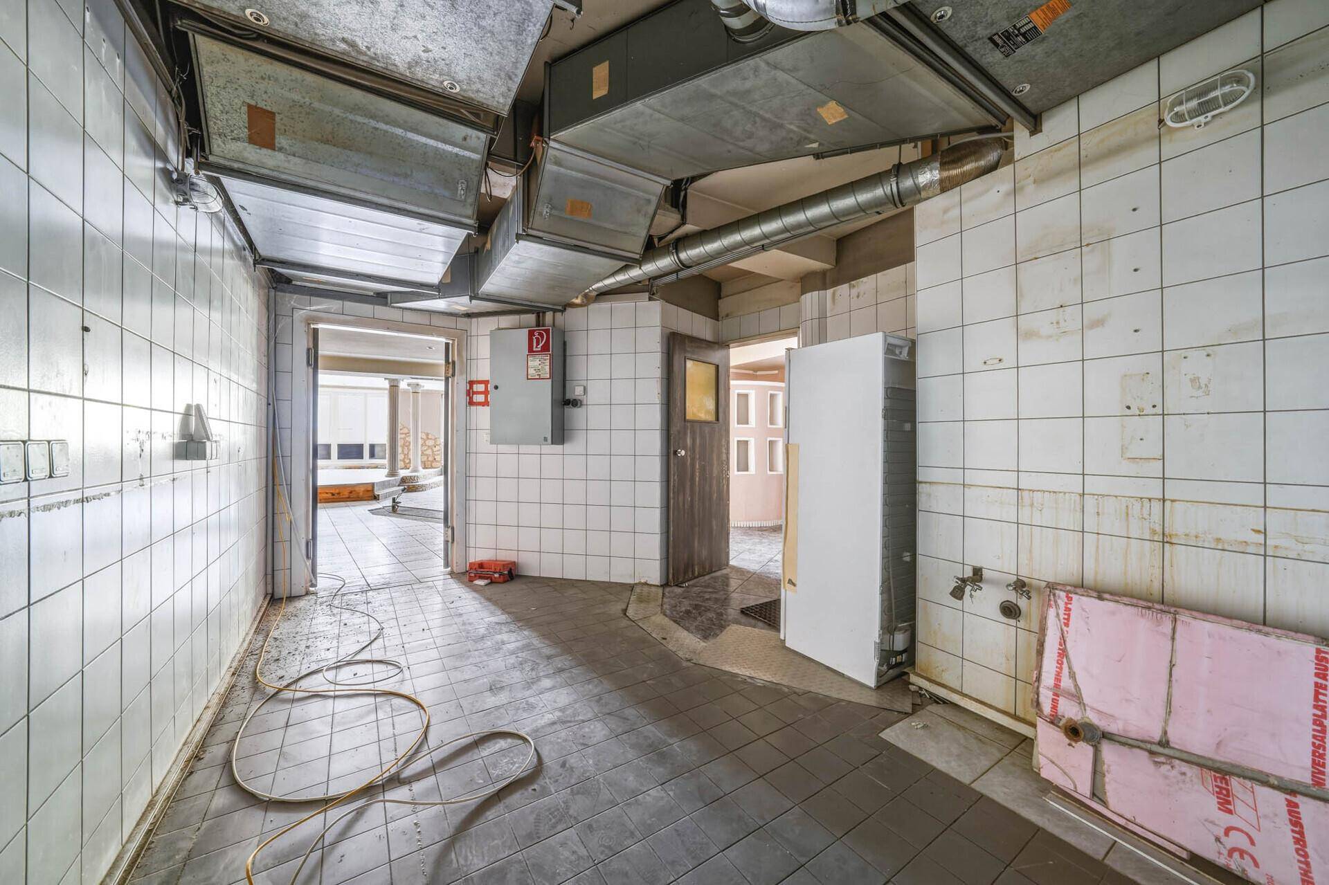 Industrielle Küche mit weißen Fliesen, sichtbaren Lüftungsrohren und einem Kühlschrank, bereit zur Nutzung.