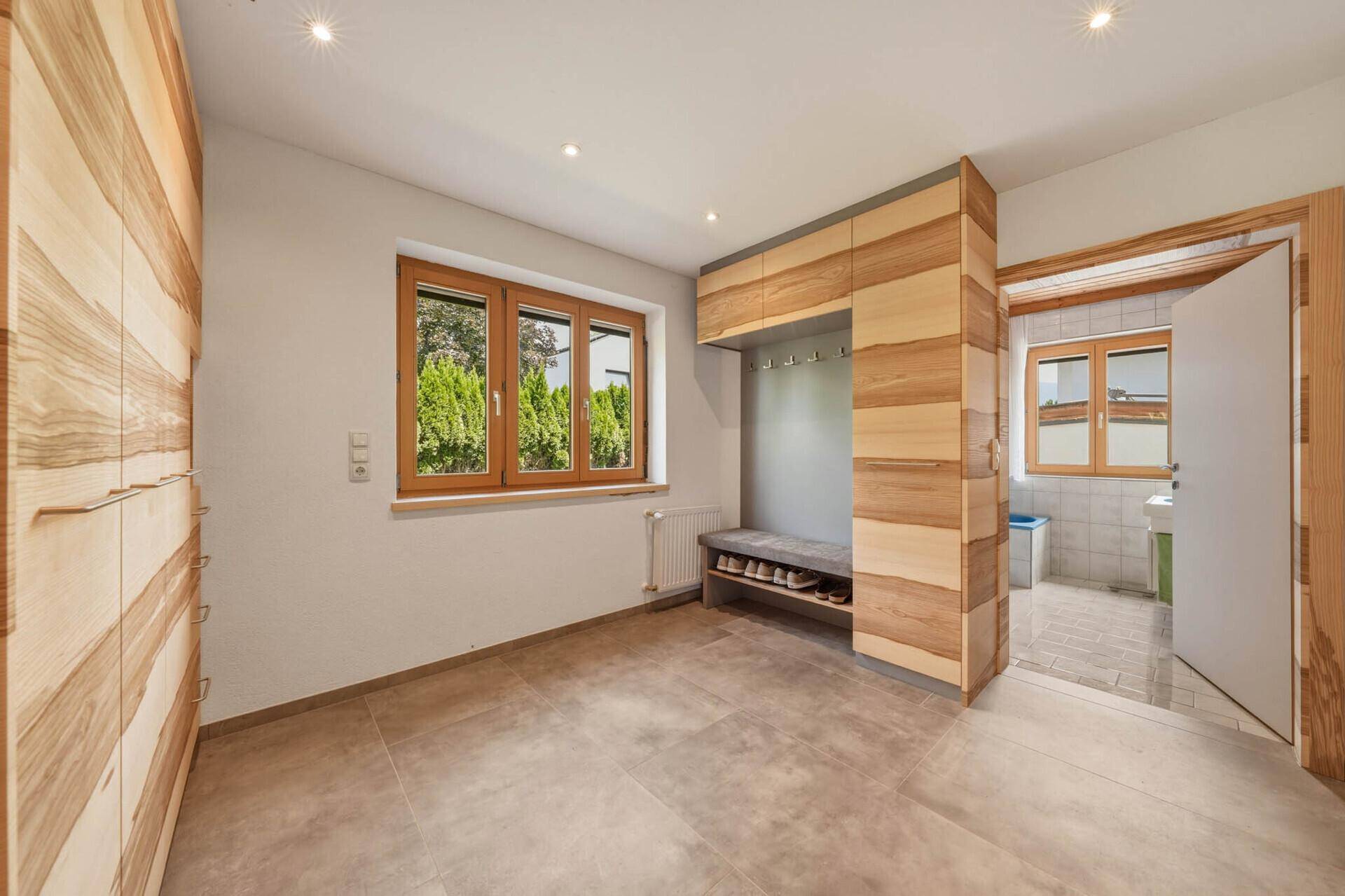 Flur mit Einbauschränken aus Holz, Fliesenboden und einem Fenster mit Blick ins Grüne.