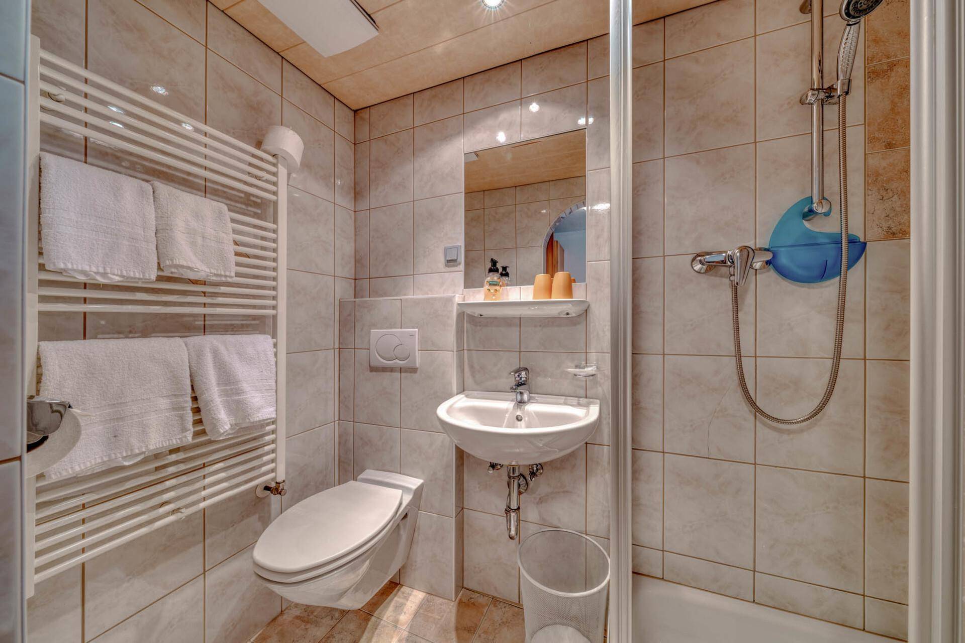 Kleines Badezimmer mit beigefarbenen Fliesen, Wandtoilette, Waschbecken und Duschbereich.