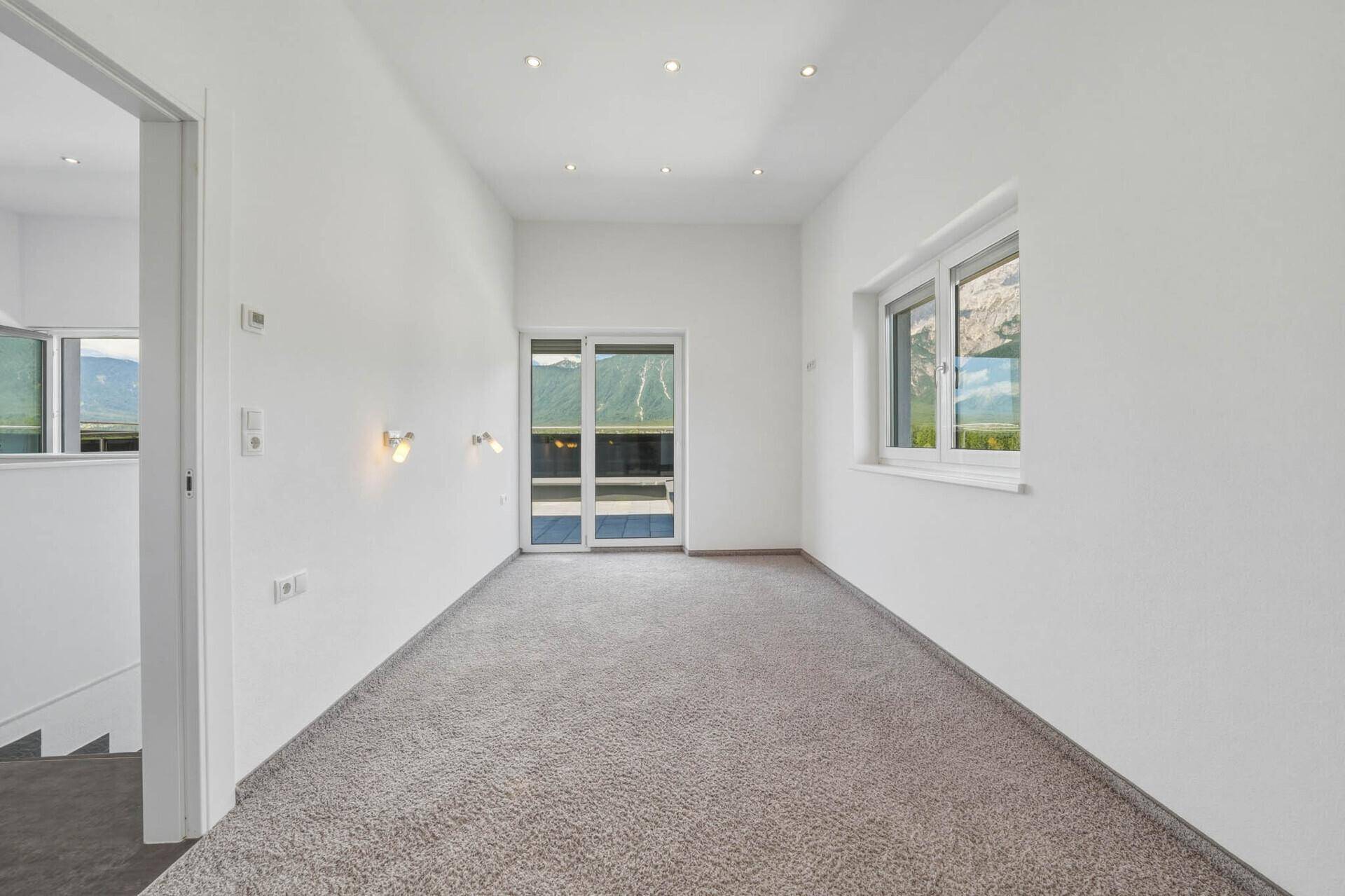 Heller Raum mit Teppichboden, Zugang zur Terrasse und Bergblick durch große Fenster.