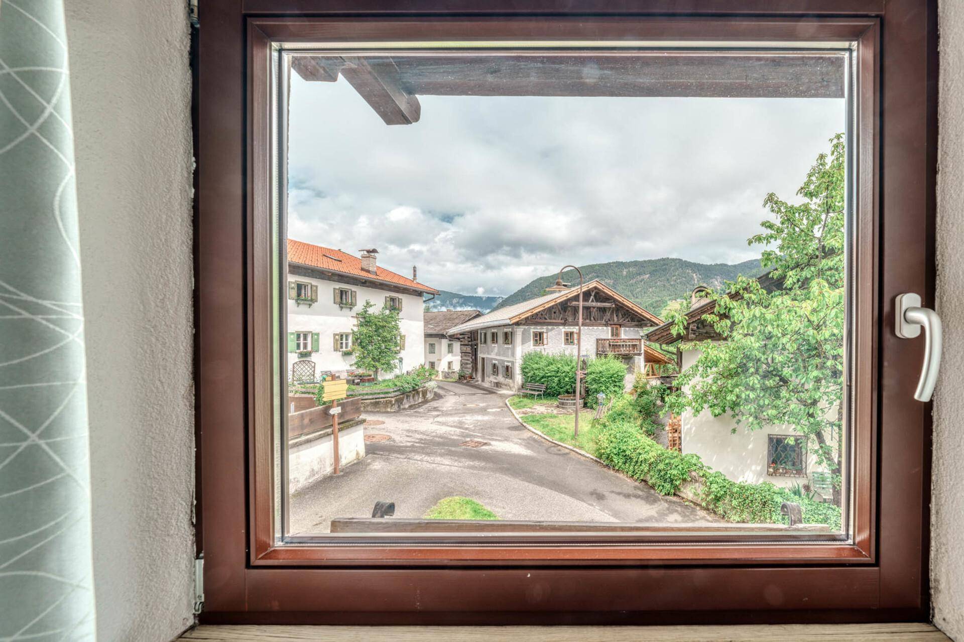 Blick aus dem Fenster auf eine ländliche Straße mit traditionellen Häusern und Bergen im Hintergrund.