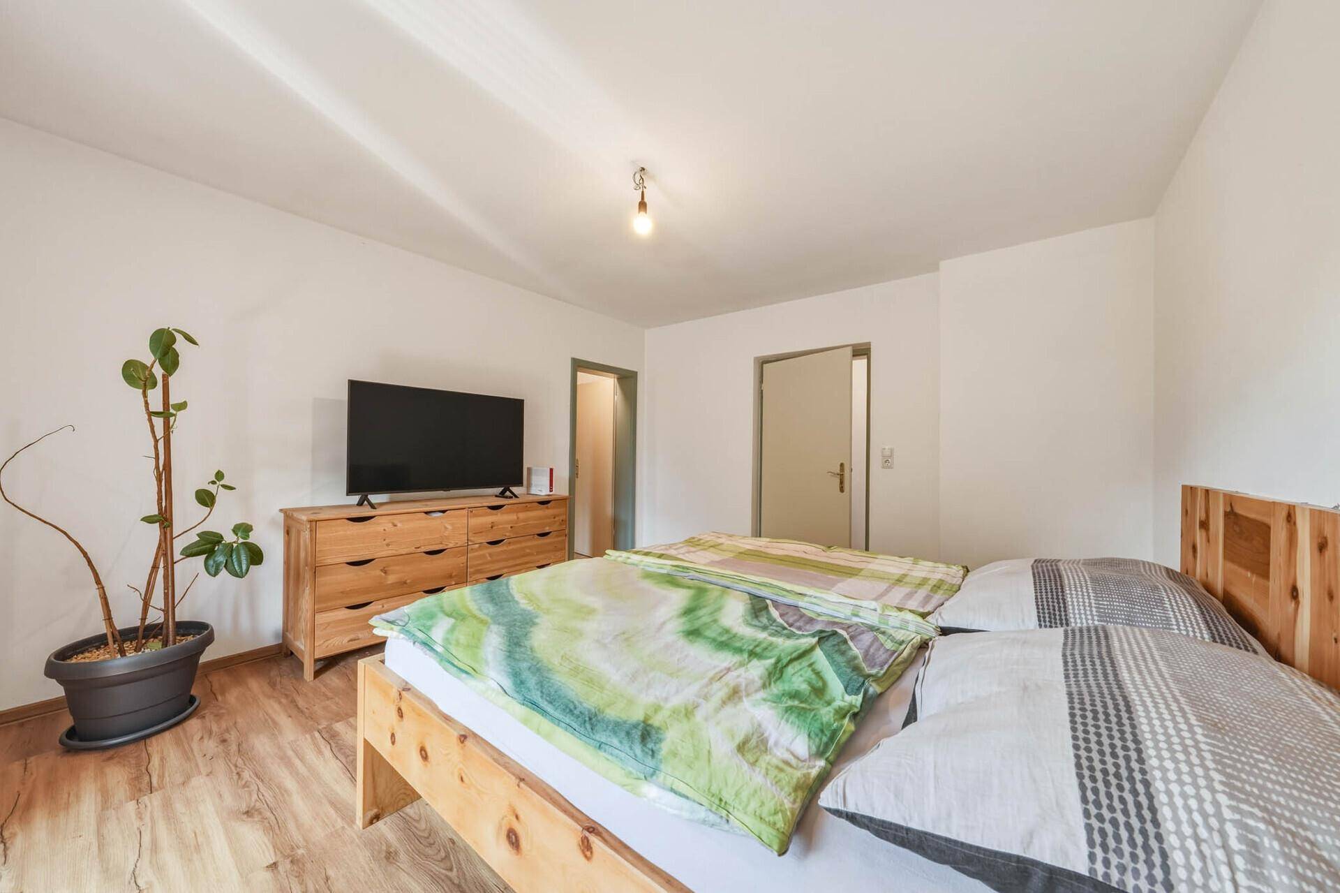 Schlafzimmer mit Holzbett, Kommode, Fernseher und Laminatboden in hellem Ambiente.