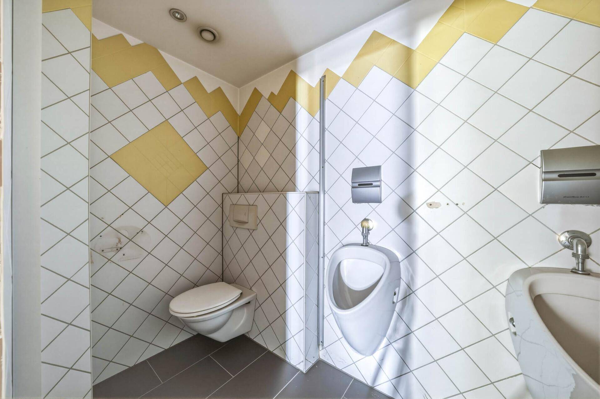 Moderne Gästetoilette mit WC, Urinal und diagonalen Fliesen in Weiß und Gelb.