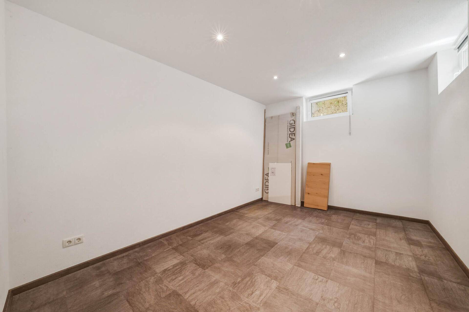 Helles, leeres Zimmer mit Fliesenboden und einem kleinen Fenster, das Tageslicht hereinlässt.