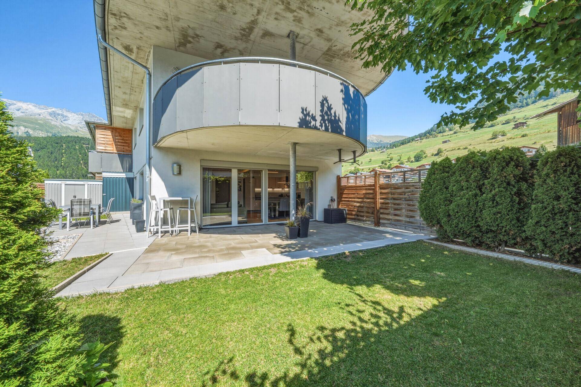 Moderne Gebäudefassade mit Terrasse, Garten und geschwungenem Balkon vor Bergkulisse.