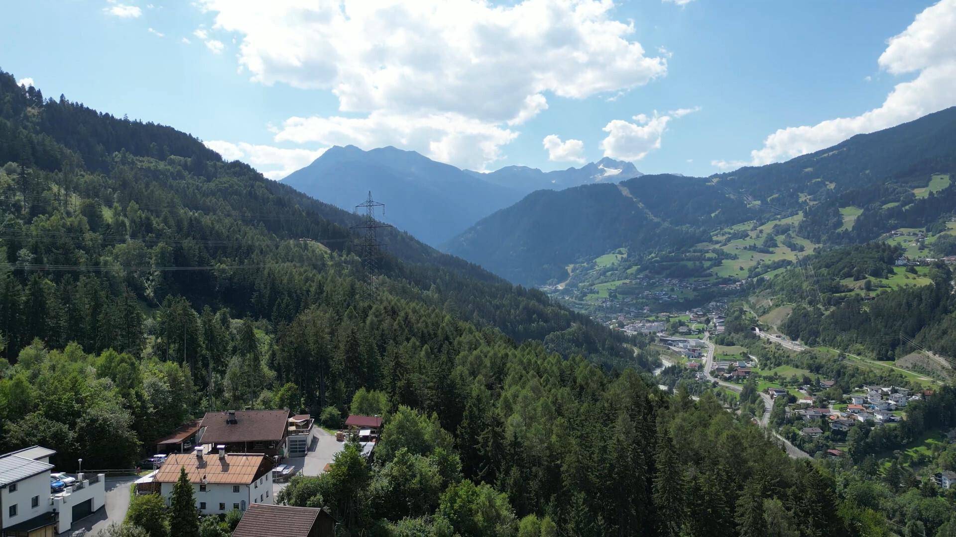 Weitläufiger Ausblick über ein bewaldetes Tal mit einem Dorf und majestätischen Bergen im Hintergrund.