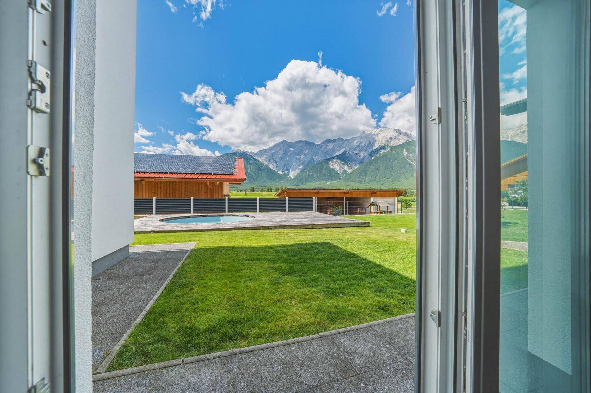 Blick aus dem Fenster auf den gepflegten Garten mit Rasenfläche, kleinem Pool und majestätischem Bergpanorama.