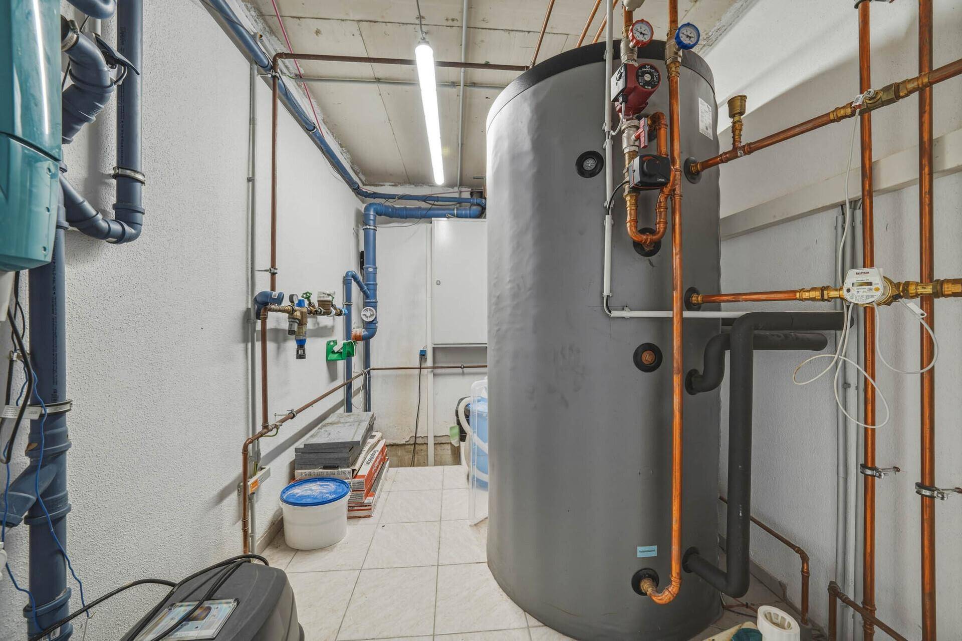 Technikraum mit großem Warmwasserspeicher, zahlreichen Rohren und funktionalem Fliesenboden.