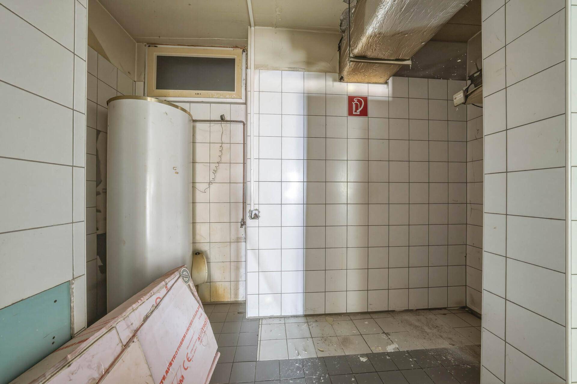 Ein gefliester Raum mit einem Warmwasserspeicher, einem Urinal und sichtbaren Lüftungsrohren an der Decke.