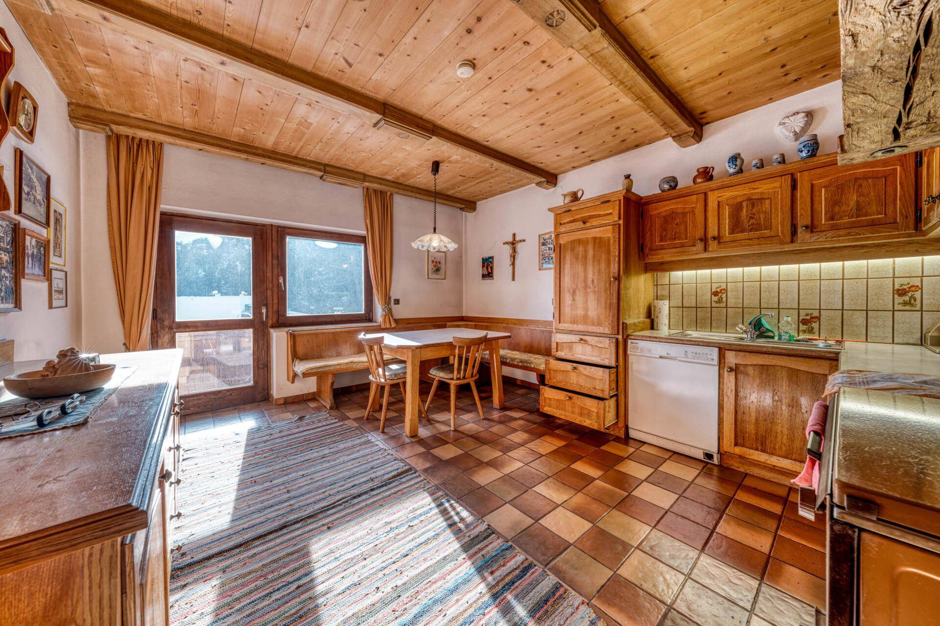 Rustikale Wohnküche mit Holzbalkendecke, Fliesenboden und Essbereich am Fenster.